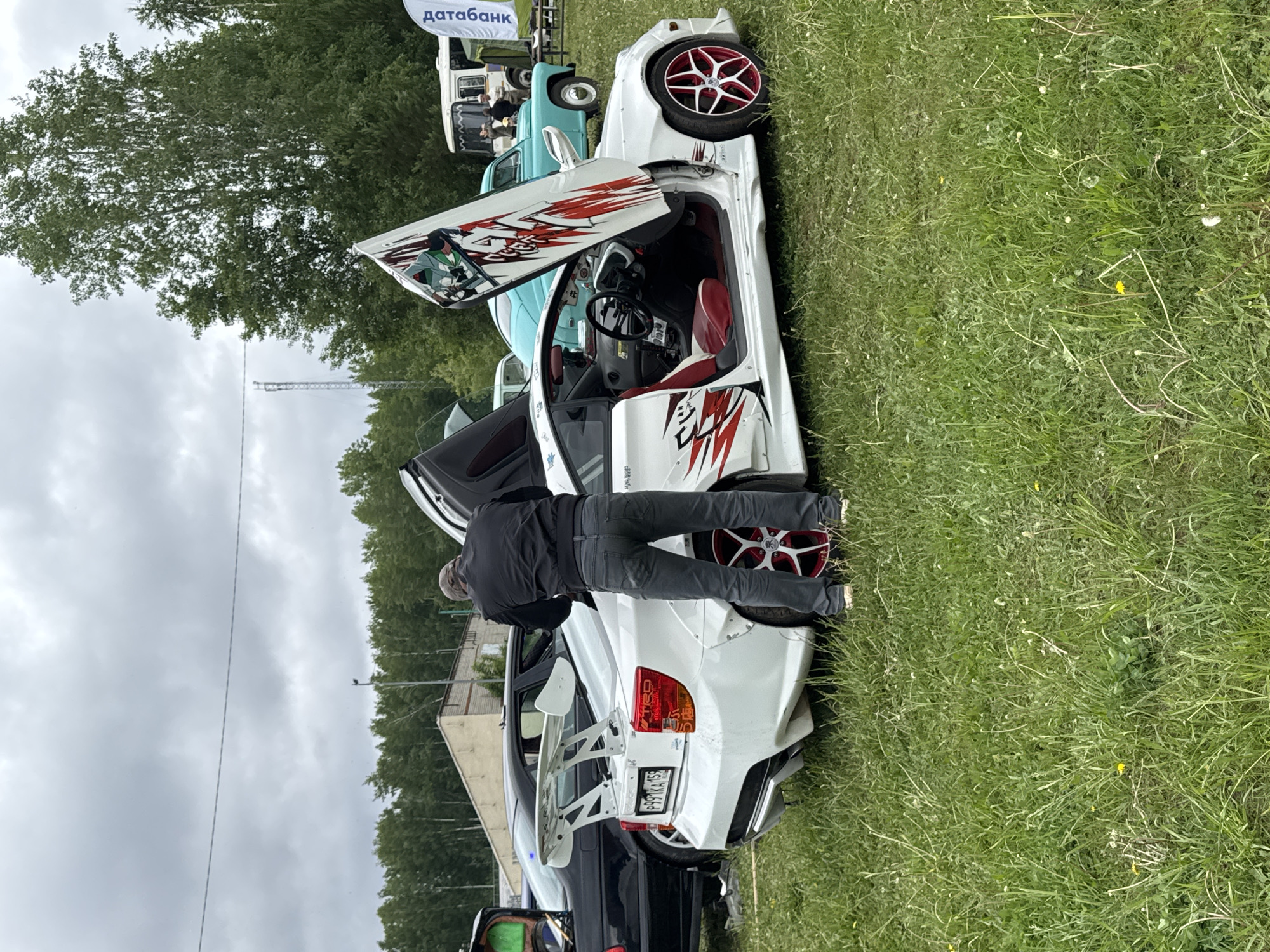 In Chaikovsky, Russia, a man in a dark jacket and gray pants is inspecting a customized white car with red and white accents. The car's doors are open, revealing a red interior. The car is parked on a grassy area. In the background, there are other vehicles and trees. The sky is overcast.
