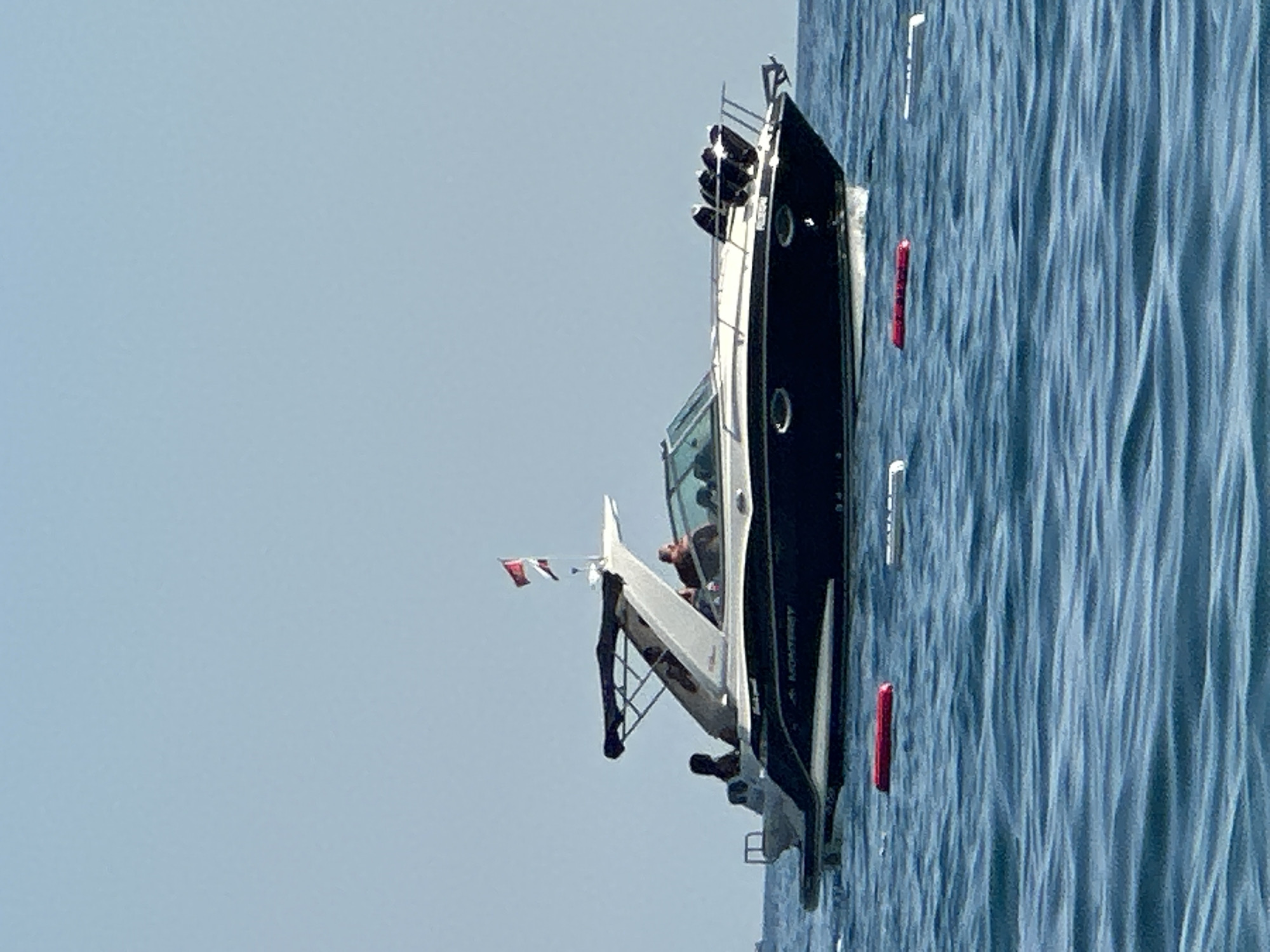 A dark blue and white Monterey boat is docked in the blue waters off the coast of Boreti, Montenegro. The boat has a canopy and two people are visible on board, one in the cockpit and another person standing at the stern. Several red and white buoys are floating in the water around the boat. The sky is clear and blue.