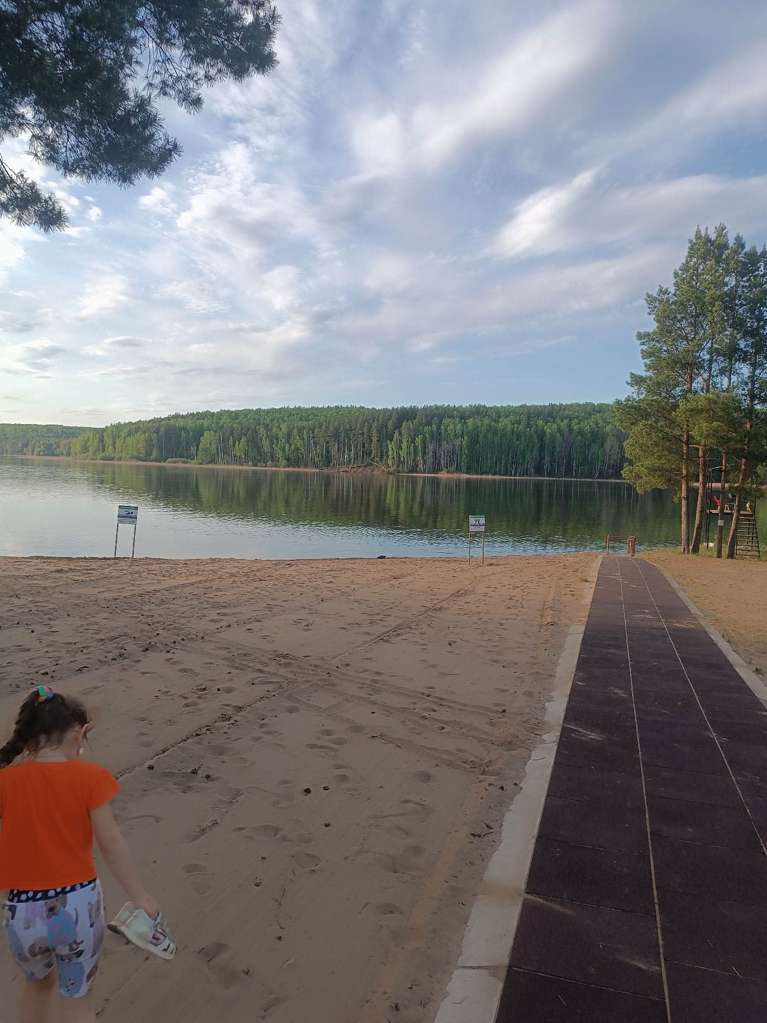 A daytime outdoor scene features a sandy beach leading to a body of water, likely a lake, fringed by a dense forest in the background. The sky is partly cloudy with a mix of blue and white hues. A young girl wearing an orange shirt and patterned shorts stands in the foreground, holding a pair of shoes. A dark-colored walkway extends from the beach towards the water, situated next to trees and a ladder structure. Signs are posted near the water's edge. Tire tracks are visible in the sand. The location appears to be in a natural setting. The specific city is not discernible from the image.