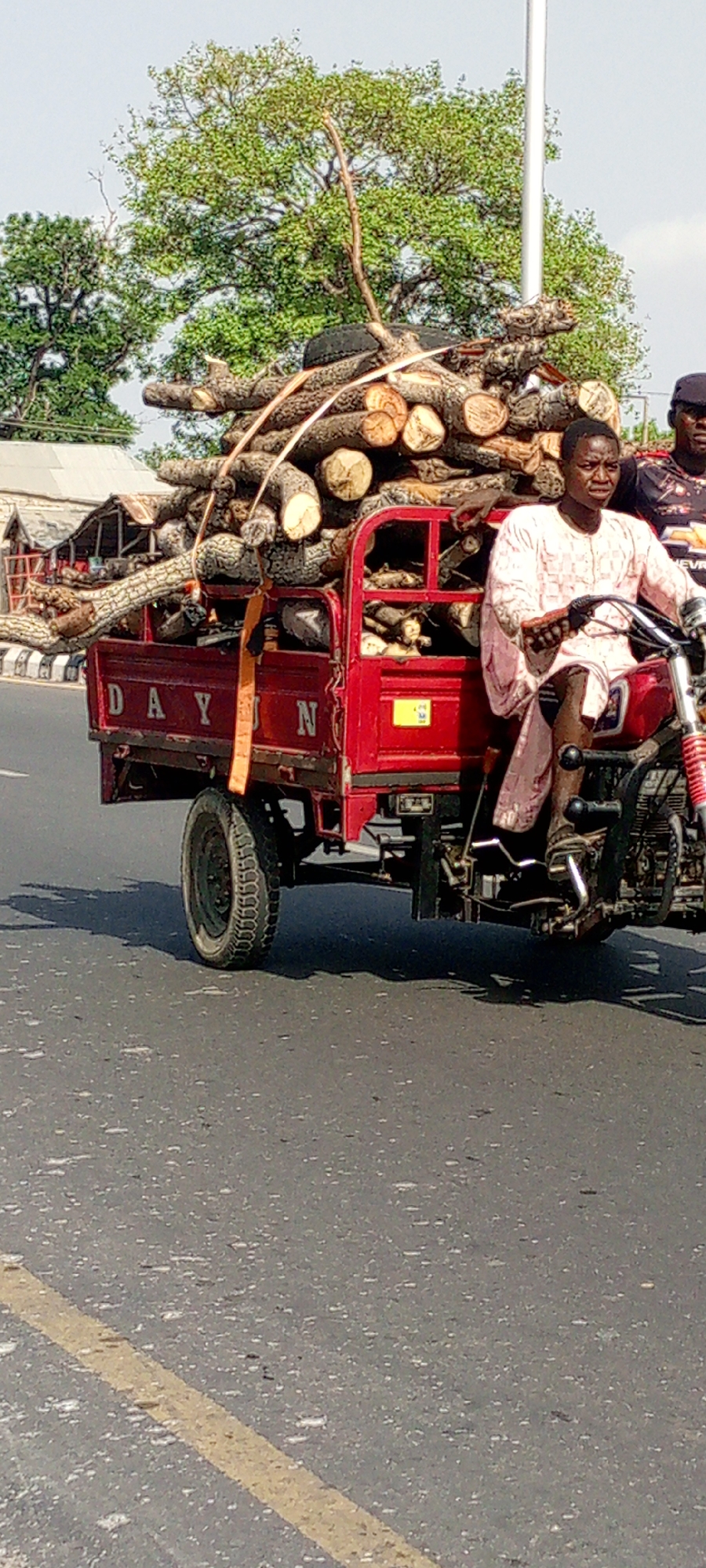 In Adankolo, Nigeria, a man operates a red DAYUN three-wheeled motorcycle heavily loaded with tree logs. The logs fill the rear bed, extending well above its sides. The man is wearing a light pink shirt and is focused on driving. Beside him is a second man wearing a black cap and a t-shirt with a Chevrolet logo. The vehicle is on a paved road. In the background, there are trees and building structures. A utility pole is also visible.