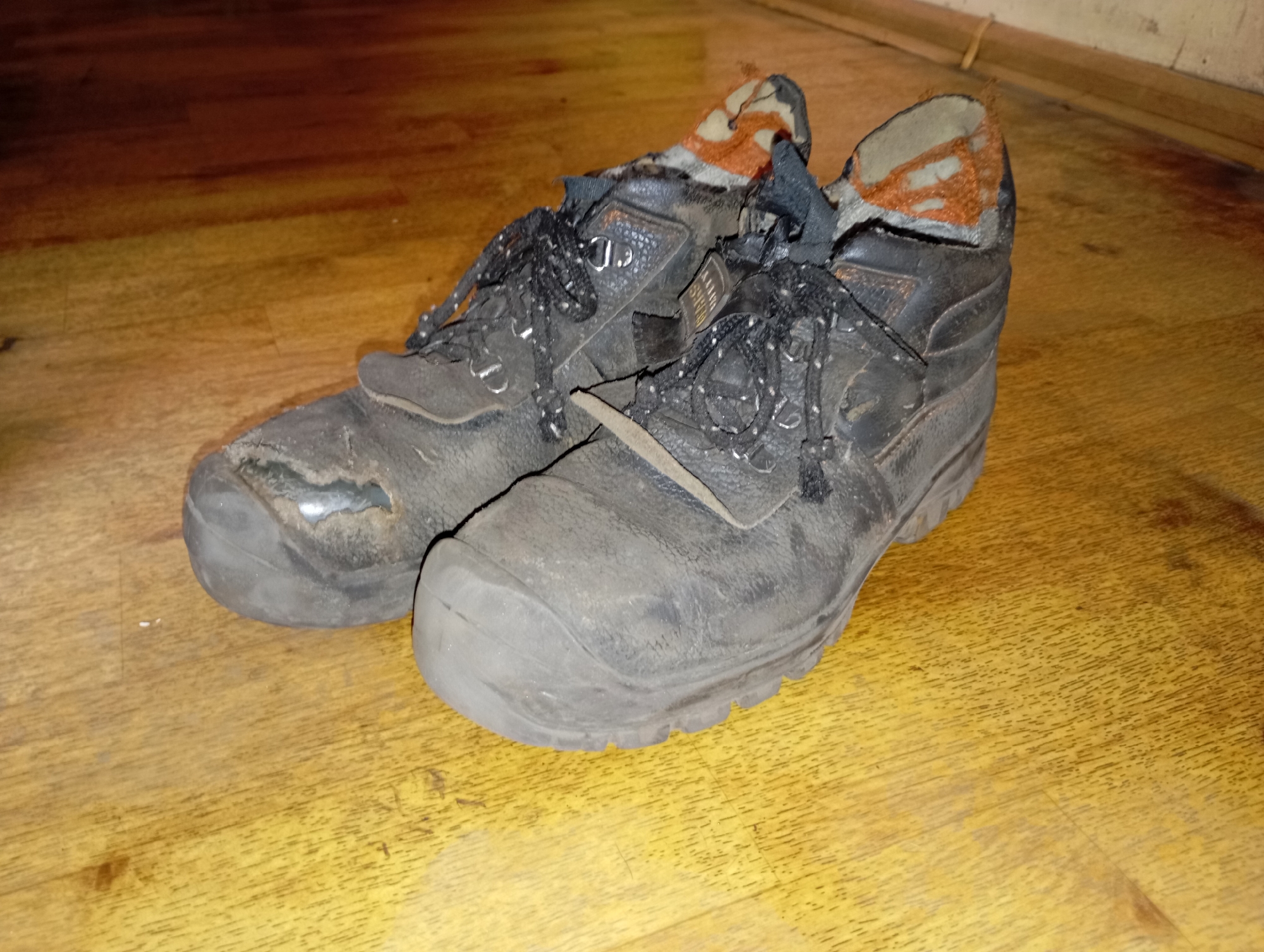 A pair of worn, black, leather work boots with orange and tan linings are placed on a light brown wooden floor. The boots are laced with black laces and show significant signs of wear, including scuffs and a tear on the toe of the left boot. The scene appears to be indoors, possibly a room in a residential building in Brocēni, Latvia, given the visible wooden flooring and baseboard. There are no people or active interactions depicted. The lighting suggests an artificial light source.