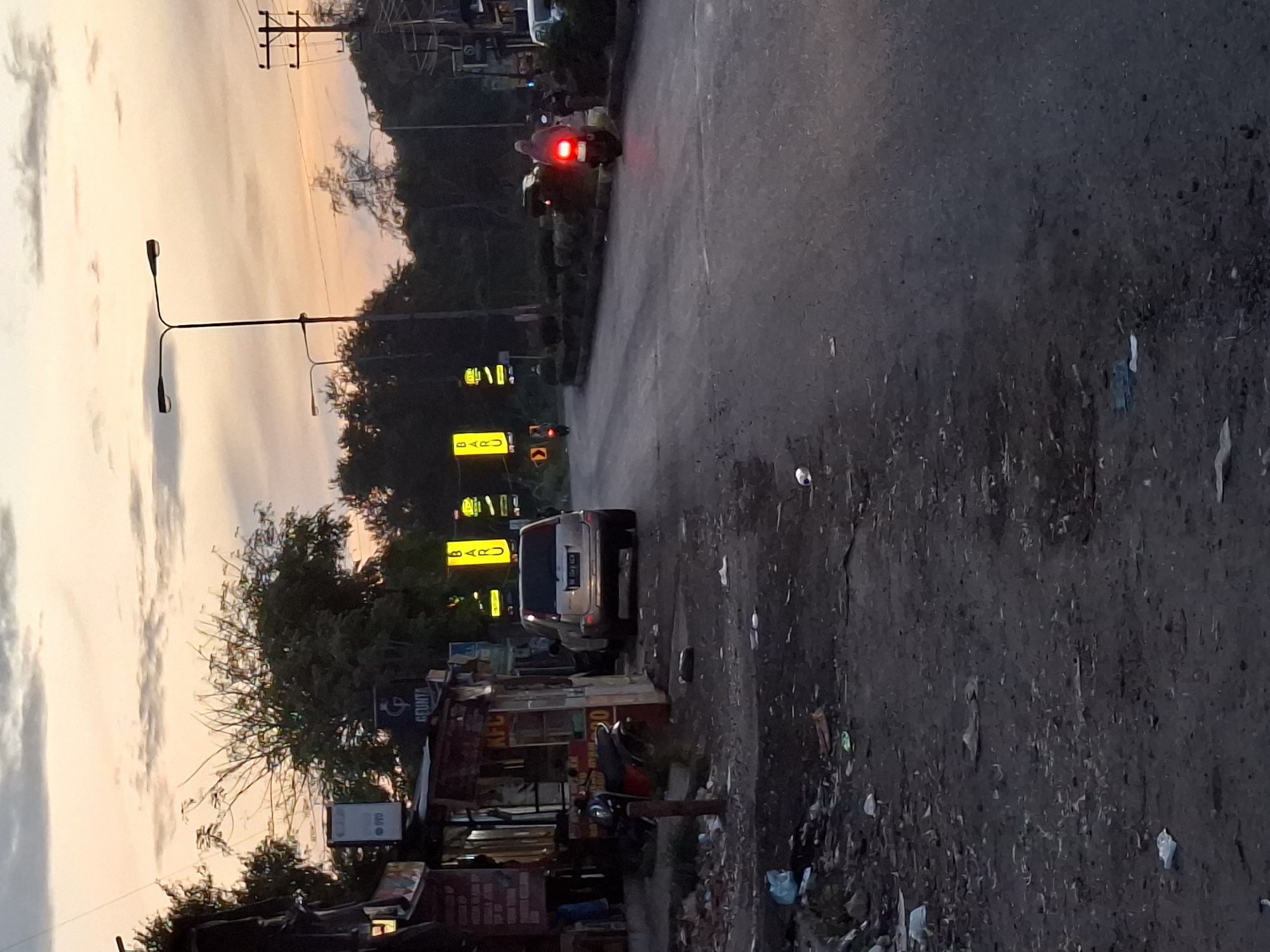 The image shows a street scene in Lhokseumawe, Indonesia at dusk. A paved road runs diagonally through the frame, with litter and debris along the shoulder. On the left side of the road, there are small shops or stalls lining the street. Signage with the word "BARU" is visible. A light-colored car is parked on the side of the road, while a motorcycle with its brake lights on is further up the road. A utility pole and streetlights are visible against the twilight sky. Trees and other vegetation are present along the roadside.