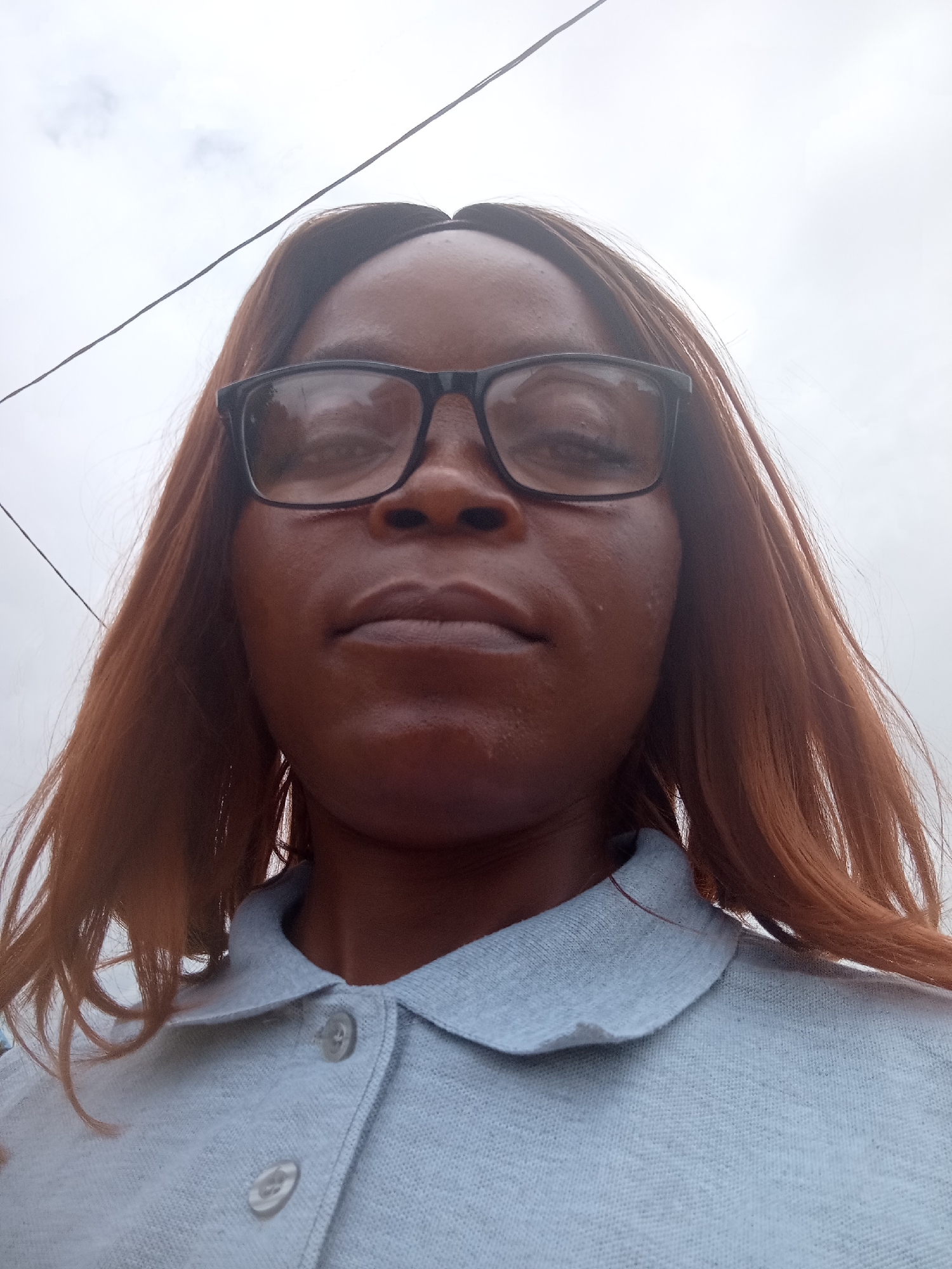 A close-up, low-angle, outdoor selfie shows a Black woman with reddish-brown hair wearing black-rimmed glasses and a light blue polo shirt. The background is an overcast sky with a thin black wire running diagonally from the top left to the top right corner. The woman's face is turned slightly upwards and towards the camera, with her mouth closed and her gaze directed slightly off-camera. A slight sheen of sweat is visible on her forehead and upper lip. The image does not contain any elements that suggest a specific location within Kason, Nigeria.