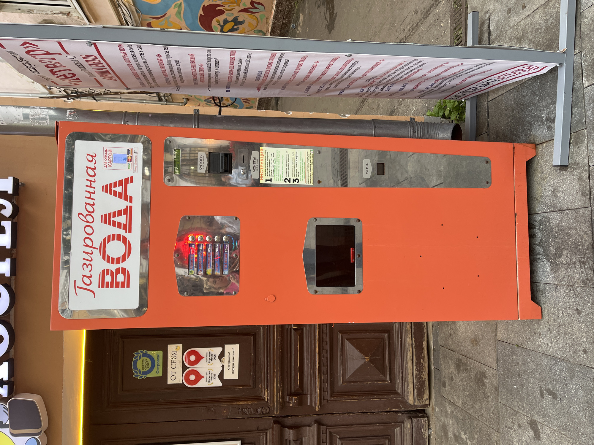 This is a vertically oriented, eye-level, medium shot of an orange water vending machine installed on a sidewalk in Nizhny Novgorod, Russia. The machine has several compartments. On the left, a large vertical sign reads "Газированная ВОДА" (Sparkling WATER) in large red Cyrillic letters. Below this sign, a smaller rectangular panel displays logos for Visa and Mastercard, indicating card payment is accepted. To the right of the water sign, there's a coin slot labeled "КУПЮРЫ" (BILLS) and a slot for coins labeled "МОНЕТЫ" (COINS). Above the coin slot is a printed instruction manual in Russian, detailing how to use the machine. Further to the right, there's a silver panel with a dark, empty screen or dispenser area, and a small button labeled "СДАЧА" (CHANGE). A string of battery-like objects, possibly for power or a display, is visible behind a transparent panel in the upper-left section of the machine's main orange body.
The vending machine is positioned in front of a dark wooden door with ornate carvings. Above the machine, a banner with Cyrillic text is attached to a metal frame. The sidewalk is paved with grey stone. Some stickers are visible on the wooden door: one is a blue circular badge with "GUESTS' CHOICE 2025" and Cyrillic text, and another is a red and white circular sticker with "ОТ СЕБЯ" (from myself) and further Cyrillic text related to good places. Another sticker warns "Осторожно! Внутри скользко!" (Caution! Slippery inside!). The overall lighting suggests it is daytime, possibly overcast. The scene appears to be in an urban environment, given the sidewalk and building facade. The presence of the vending machine and the door suggest a commercial or public space. There are no people visible in the frame.