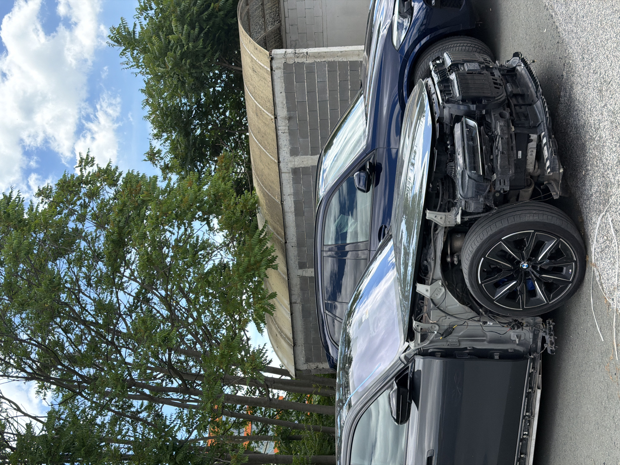 The image captures an outdoor scene in Burgas, Bulgaria, featuring two cars parked in front of a building with a curved roof. The car in the foreground is a dark gray sedan, heavily damaged at the front, with its hood, bumper, and grille removed, exposing the internal components. The wheel well is visible, showcasing a black rim with a silver accent. The side of the car shows damage along the lower body panel. In the background, a dark blue SUV is parked, appearing intact. Behind the vehicles, a building constructed with grey concrete blocks and a weathered, arched roof structure is visible. Lush green trees with dense foliage are present on the left and behind the building, with a blue sky with scattered white clouds overhead. The ground is paved with asphalt. There are no people visible in the frame.