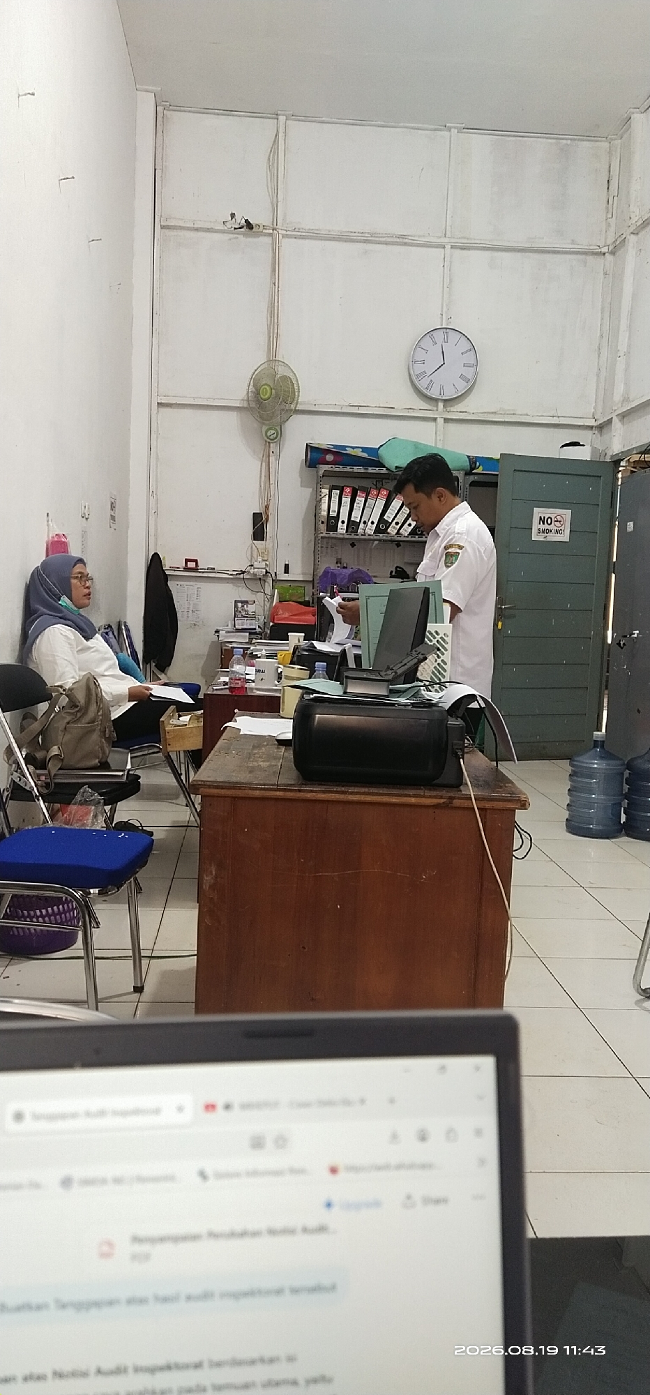 The image depicts an indoor scene, likely an office in Tamiang Layang, Indonesia. Two individuals are present. A woman wearing a hijab and glasses is seated on a chair, looking towards the right. A man in a white uniform shirt with a green emblem on the left breast is standing and looking down at papers he is holding. He appears to be in the process of reviewing documents.
The environment is cluttered with office supplies. A wooden desk in the foreground holds a black printer and various papers. A shelf unit filled with binders and files stands against the wall behind the standing man. A circular clock is visible on the wall. A green door with a "NO SMOKING" sign is to the right of the shelf. Water cooler jugs are stacked on the floor near the door. A wall fan is mounted on the wall. The floor is tiled. The overall impression is of a functional but somewhat disorganized office space. The foreground also shows a portion of a laptop screen displaying what appears to be a web page. A timestamp in the lower right corner reads "2026.08.19 11:43".