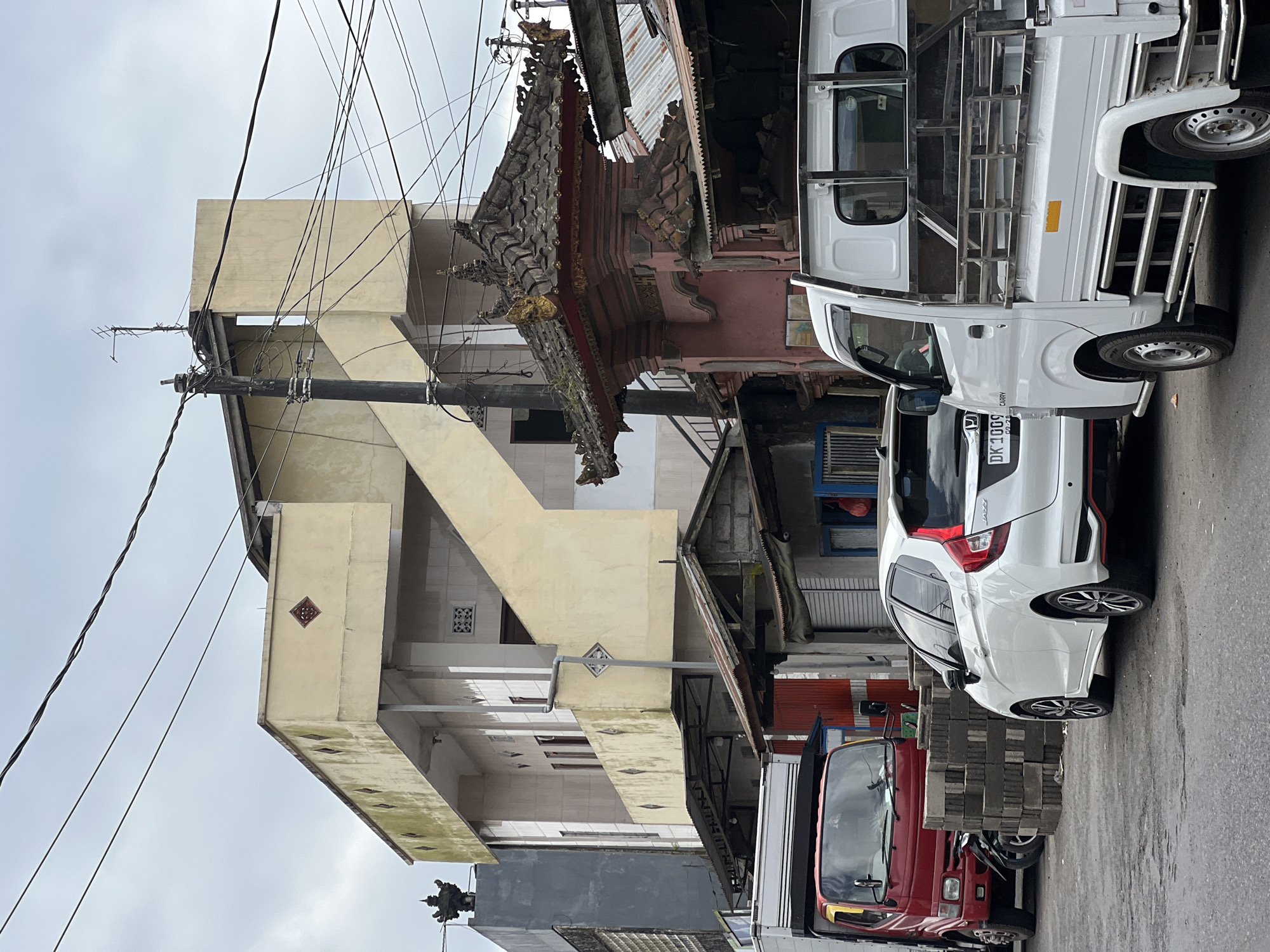 In Kintamani, Indonesia, a street scene unfolds under an overcast sky. A red delivery truck is parked on the left, partially obscured by a stack of grey concrete blocks and a motorcycle. To its right, a white hatchback car faces away from the camera, its rear visible. Beyond the vehicles, a multi-story building with cream-colored facade and green moss stains rises, featuring an external staircase. In the foreground, a white utility truck is parked, its bed equipped with metal railings. Several power lines crisscross the sky. Behind the utility truck, a traditional Balinese temple gate, featuring intricate carvings and a tiered roof, is visible. The scene depicts an urban or semi-urban environment, likely a commercial or residential area, with elements of local culture present.