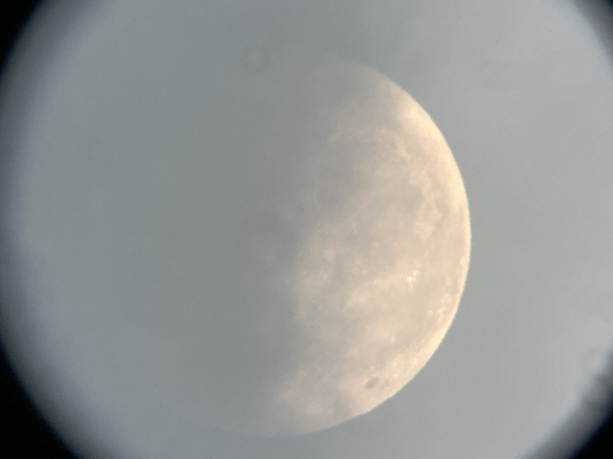 The image is a close-up, slightly out-of-focus photograph of the moon viewed through a telescope or similar optical instrument. A portion of the moon is visible, illuminated and displaying some surface texture with darker patches. The sky surrounding the moon is a hazy gray. The photograph is framed by a dark, circular border, indicating the edge of the lens or eyepiece. There is no discernible information about the city of Lyubertsy, Russia, nor any people, actions, or interactions.