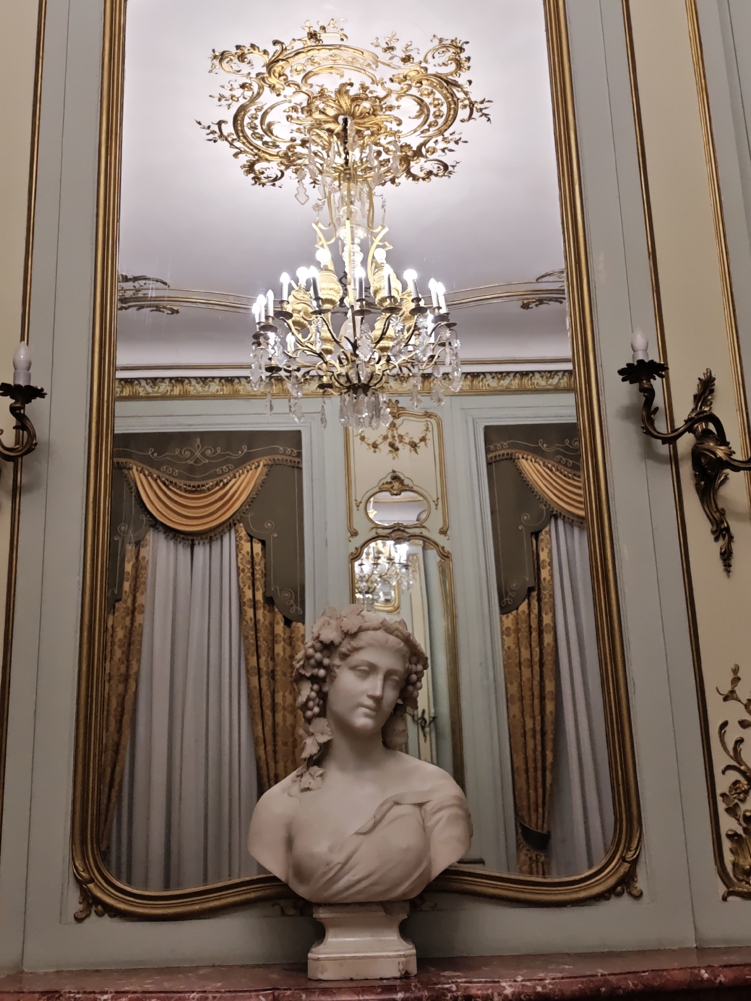 The image depicts an ornate interior scene, likely a historical building, in Lviv, Ukraine. In the foreground, a white marble bust of a woman with grapes in her hair rests on a marble surface. Behind the bust, a large, gilded mirror reflects the scene. The mirror shows a chandelier with crystal elements and golden accents hanging from a decorated ceiling. The reflection also reveals draped curtains in shades of gold and grey, and a section of wall with ornate gold trim. The room appears to be decorated in a baroque or rococo style, with elaborate moldings and fixtures. Two wall-mounted sconces with candle-like bulbs are visible on either side of the mirror frame, adding to the opulent atmosphere. The overall impression is one of grandeur and historical richness.