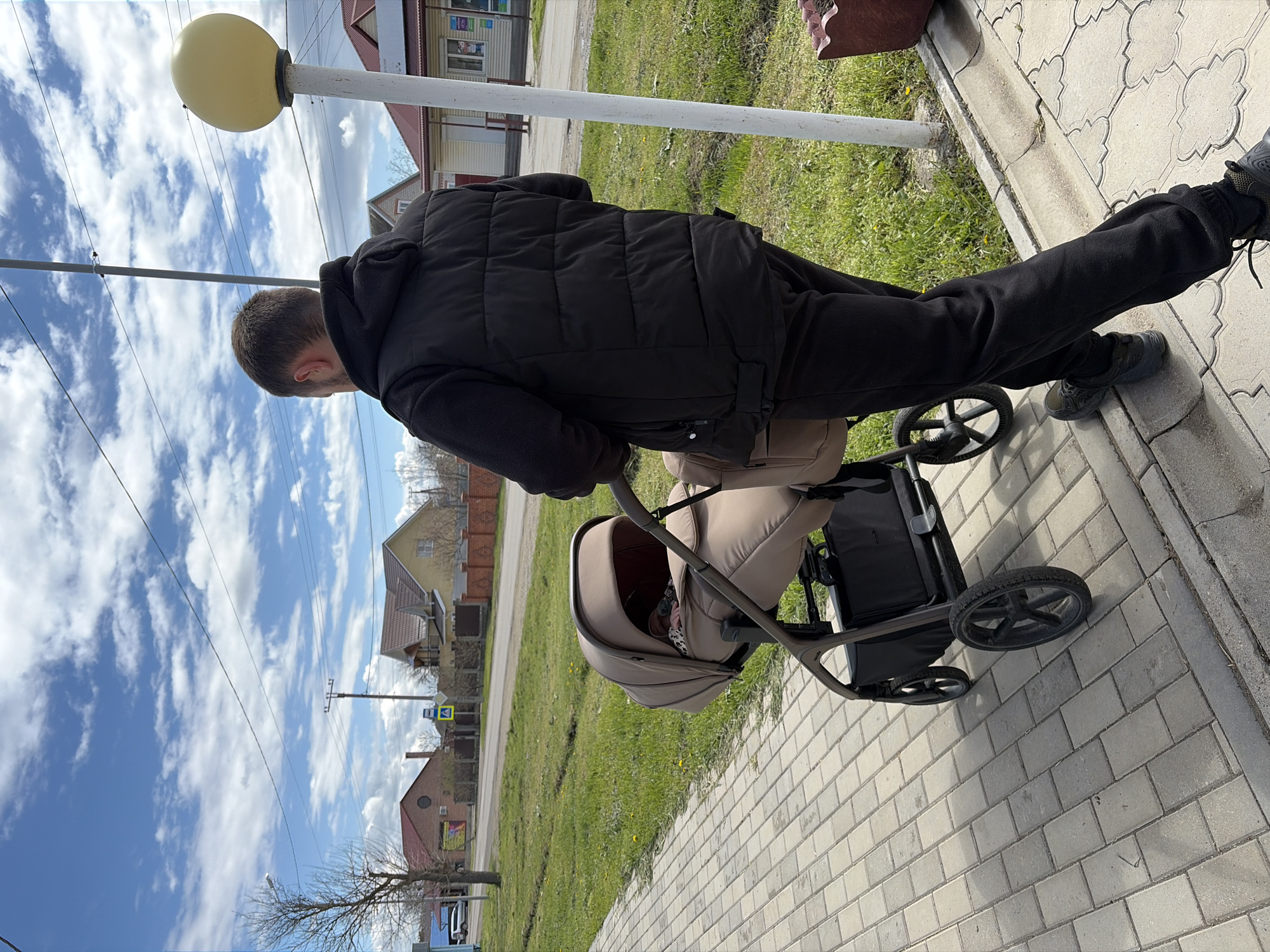A person in a black jacket and pants is pushing a stroller on a brick sidewalk. To the person's right is a curb with grass and a white post supporting a globe light. In the background are houses and a partly cloudy blue sky. The location is the city of Unknown, Unknown.