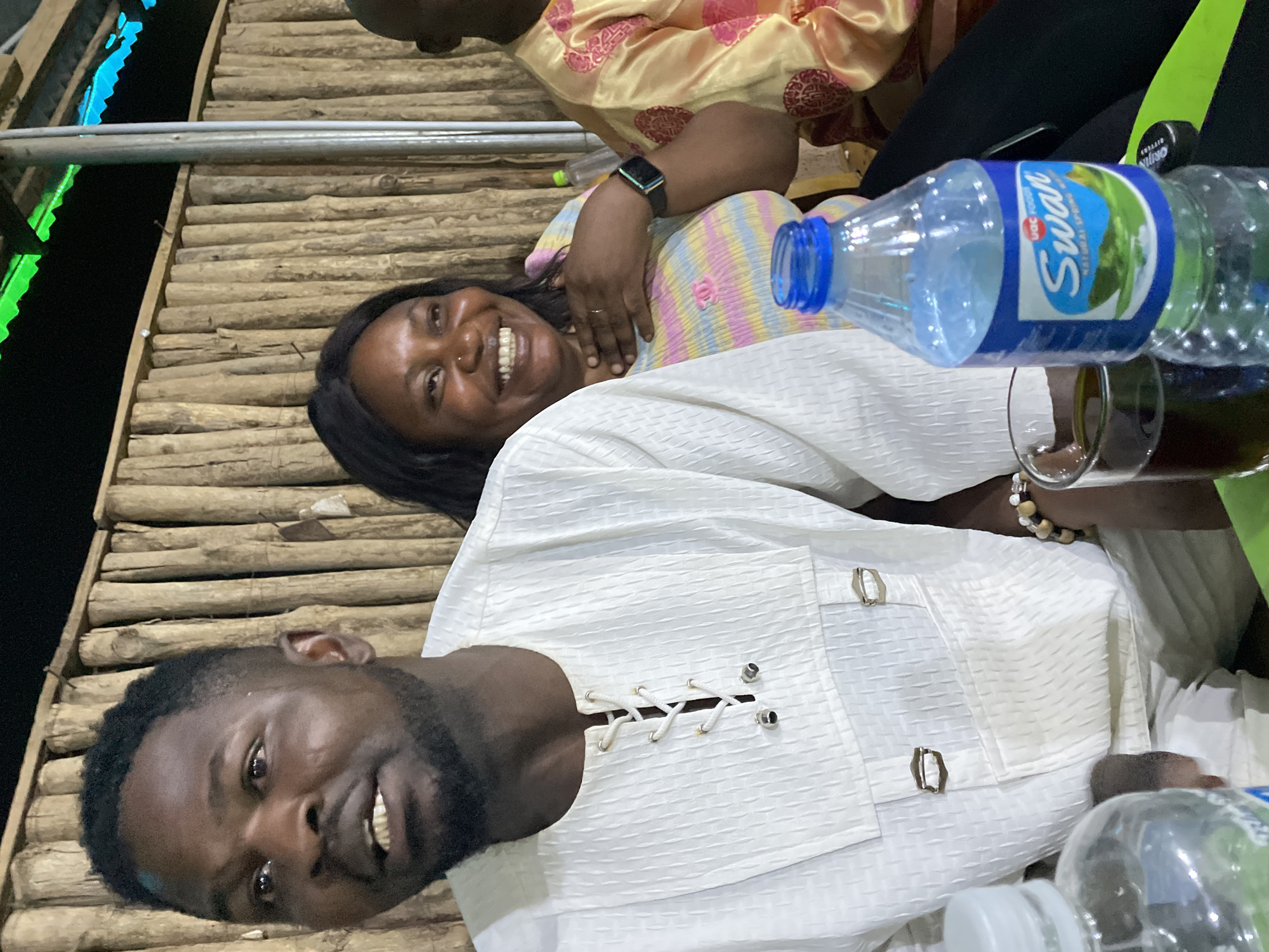 The image, originally rotated clockwise, captures a cheerful social moment, likely in Jos, Nigeria, during the evening or night. Two adults are prominently featured, smiling broadly and looking towards the camera.
In the foreground, a man with dark hair and a beard is wearing a white textured garment that features decorative lacing and metallic ring-like clasps on the chest. Behind him, a woman with dark hair also smiles widely. She is dressed in a colorful striped top, featuring shades of pink, yellow, blue, and white, and wears an Apple Watch on her left wrist, with a ring visible on her finger. A third person, wearing a yellow patterned garment, is partially visible in the background.
On the surface before them, a plastic bottle of "Swan Natural Spring Water" with a blue label is visible, alongside a glass containing a dark beverage. The subjects appear to be seated on or next to a rustic platform constructed from horizontal wooden logs or bamboo. The background is dark, illuminated by subtle hints of vibrant green and blue artificial lighting, reinforcing the nighttime setting. Both the man and woman exude happiness and contentment, suggesting a pleasant gathering or interaction. Visible text on the water bottle includes "Swan," "UAC FOODS," and "Natural Spring Water." The man also wears a beaded bracelet on his wrist.