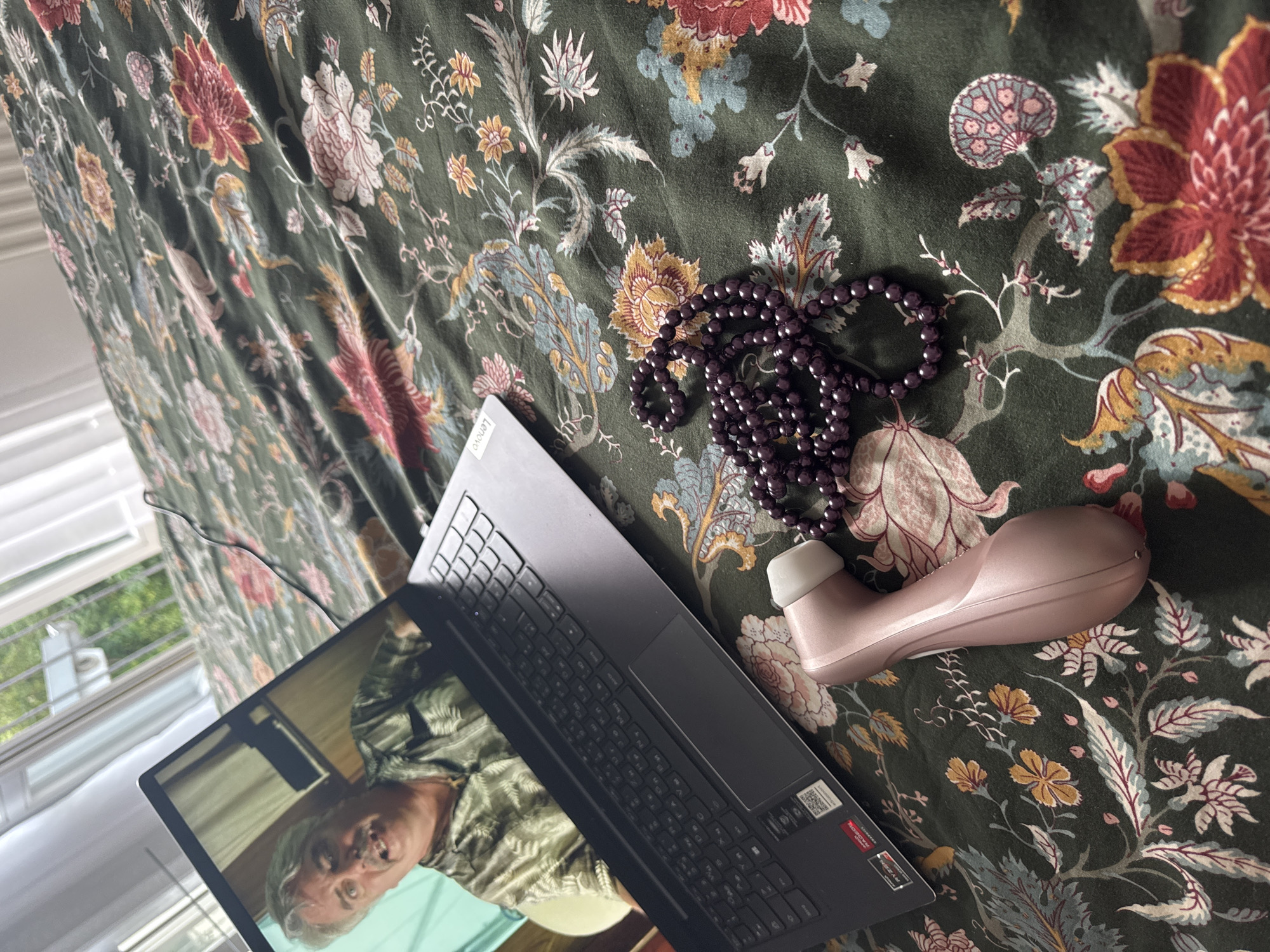 This media file depicts a flat lay of a laptop, a string of dark purple beads, and a pink massager on a dark green floral-patterned bedspread. The laptop is open, displaying a video call with an older person who appears to be a man with graying hair and wearing a green shirt. The scene is indoors, with a window visible in the background, suggesting natural light. The overall impression is a relaxed, personal setting, possibly a bedroom. There are no explicit cues about the time of day or weather, though the natural light hints at daytime. The location is specified as Höhenkirchen-Siegertsbrunn, Germany. The visible text on the laptop is a "Lenovo" logo and a "AMD Ryzen" sticker.