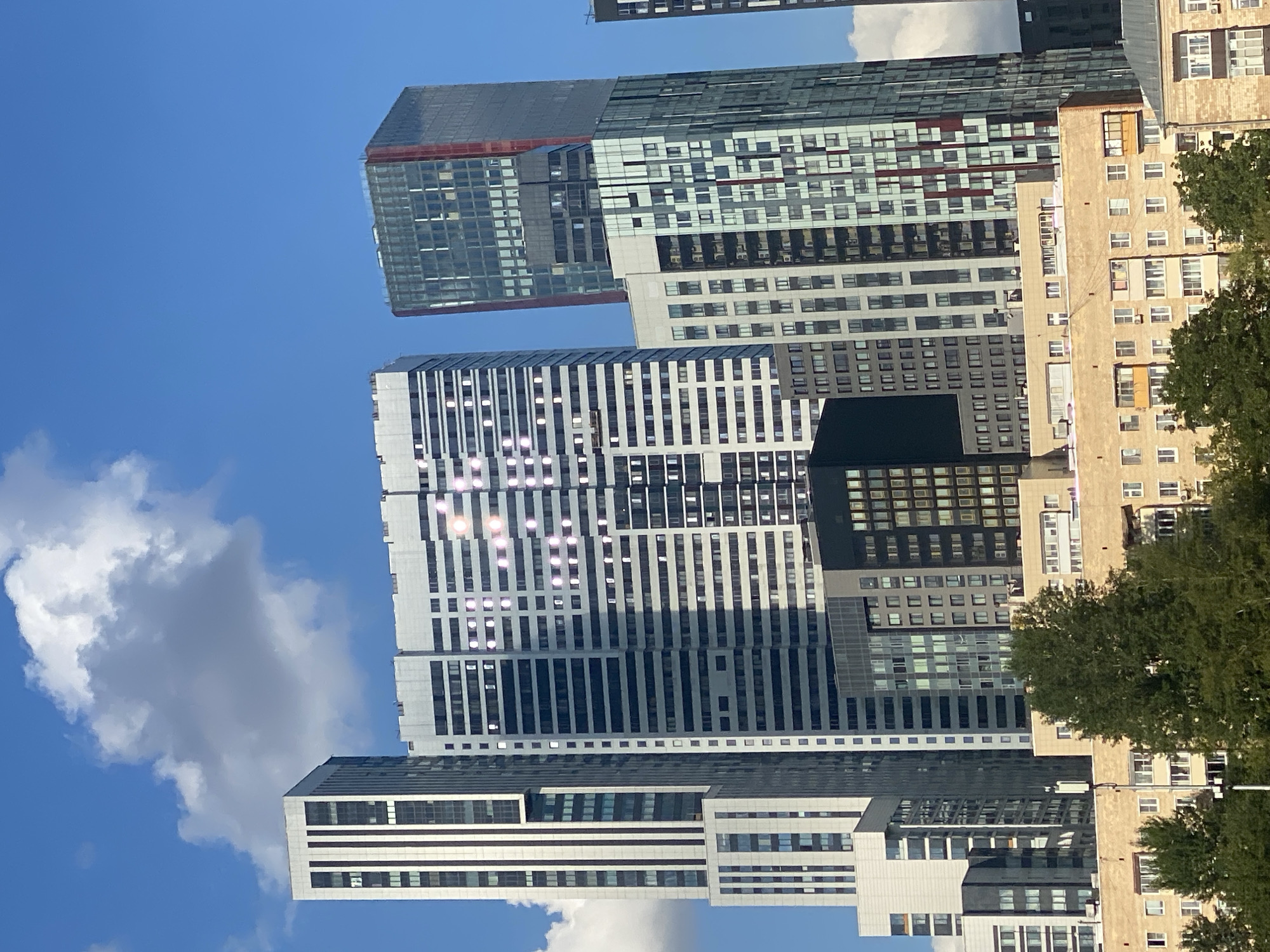 The image shows a cluster of modern skyscrapers against a clear blue sky with scattered white clouds. The buildings are of varying heights and architectural styles, with large glass facades that reflect the sunlight. One prominent building in the center has a pattern of lights visible on its side, forming a bright, abstract shape. In the foreground, lower-rise apartment buildings are visible, with trees lining the street. The overall scene depicts an urban landscape.