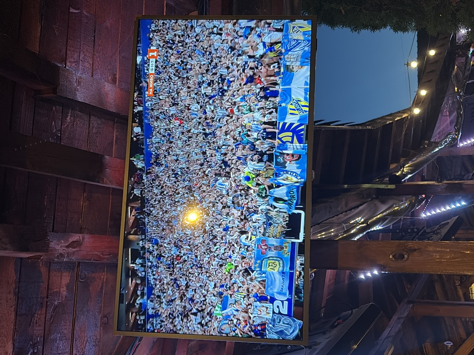 The image shows a large television screen displaying a crowded stadium filled with fans wearing white and blue striped jerseys, likely watching a soccer match. The screen has a banner at the top that reads "НА ЖИВО" (Live) and a score of "1". Below the screen, there are banners with logos, including one that says "AFA" and "ATLANTA". The TV is mounted under a wooden ceiling with exposed beams. In the background, there is a clear blue sky and some outdoor lighting. The setting appears to be an outdoor seating area, possibly a bar or restaurant, in Sofia, Bulgaria.