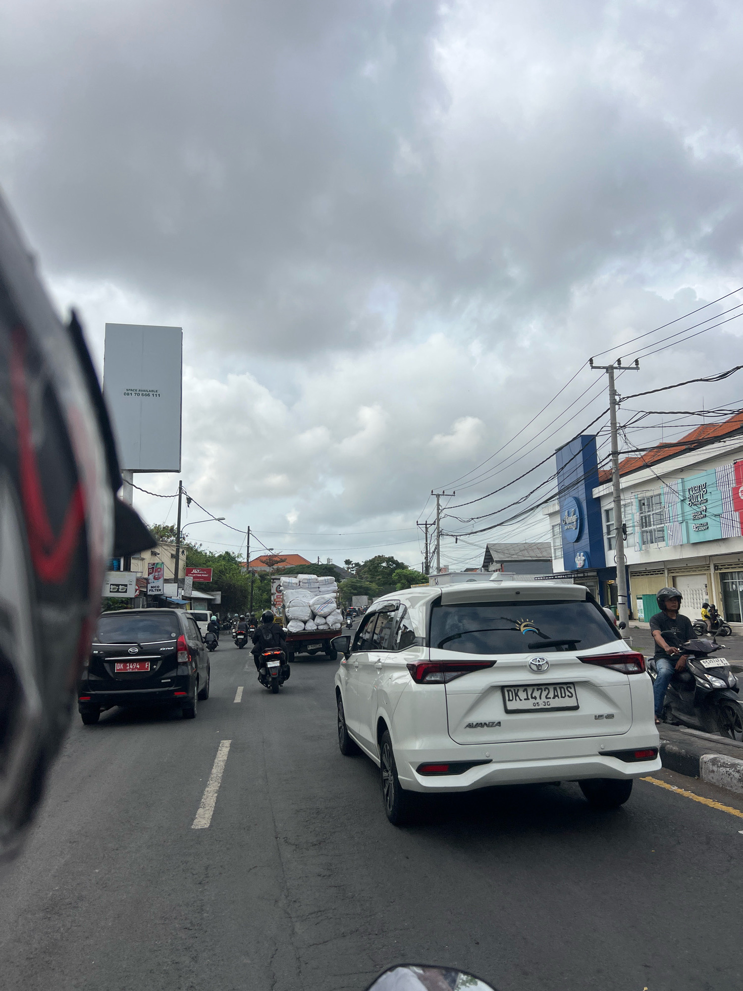 The image depicts a street scene in Denpasar, Indonesia, on a cloudy day. A dark-colored SUV is in the left lane of a multi-lane road, and a white SUV is in the right lane. Both vehicles have license plates. Several motorbikes, some with riders, are also present on the road. In the background, there are buildings with signs, power lines, and a truck loaded with large white bags. The foreground shows a portion of a helmet and a motorbike's rearview mirror, suggesting the image was taken from a moving motorbike. The road surface is asphalt with white lane markings.