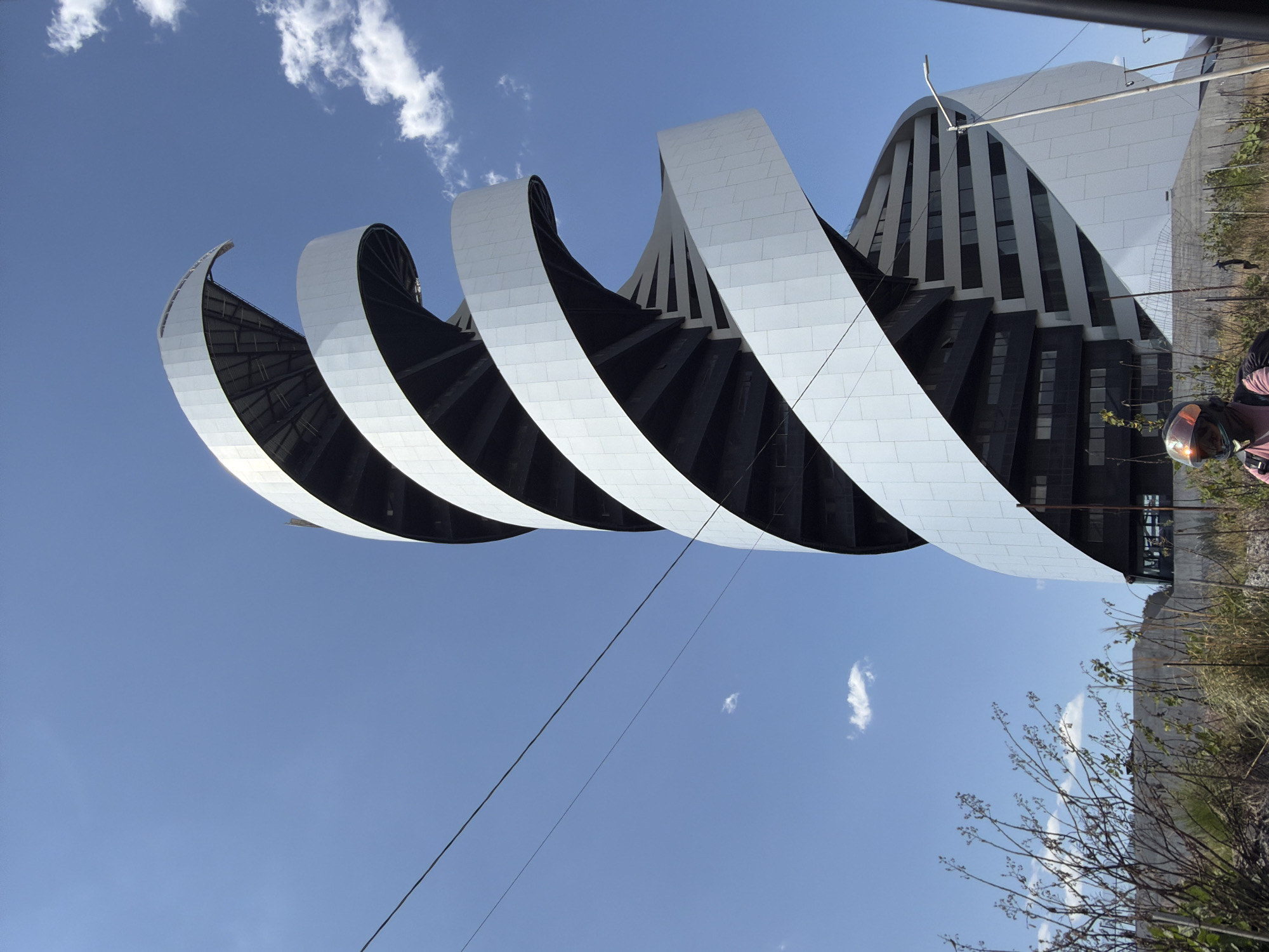 The image shows a unique architectural structure in San Bernardino Tlaxcalancingo, Mexico. The building features a series of curved, white-tiled elements that appear to spiral around a darker, presumably interior structure. The sky is a clear, bright blue with scattered clouds. The building appears to blend into a more typical building on the right side of the image. There is a figure with a helmet on. The foreground contains vegetation and an electrical line stretching across the sky.