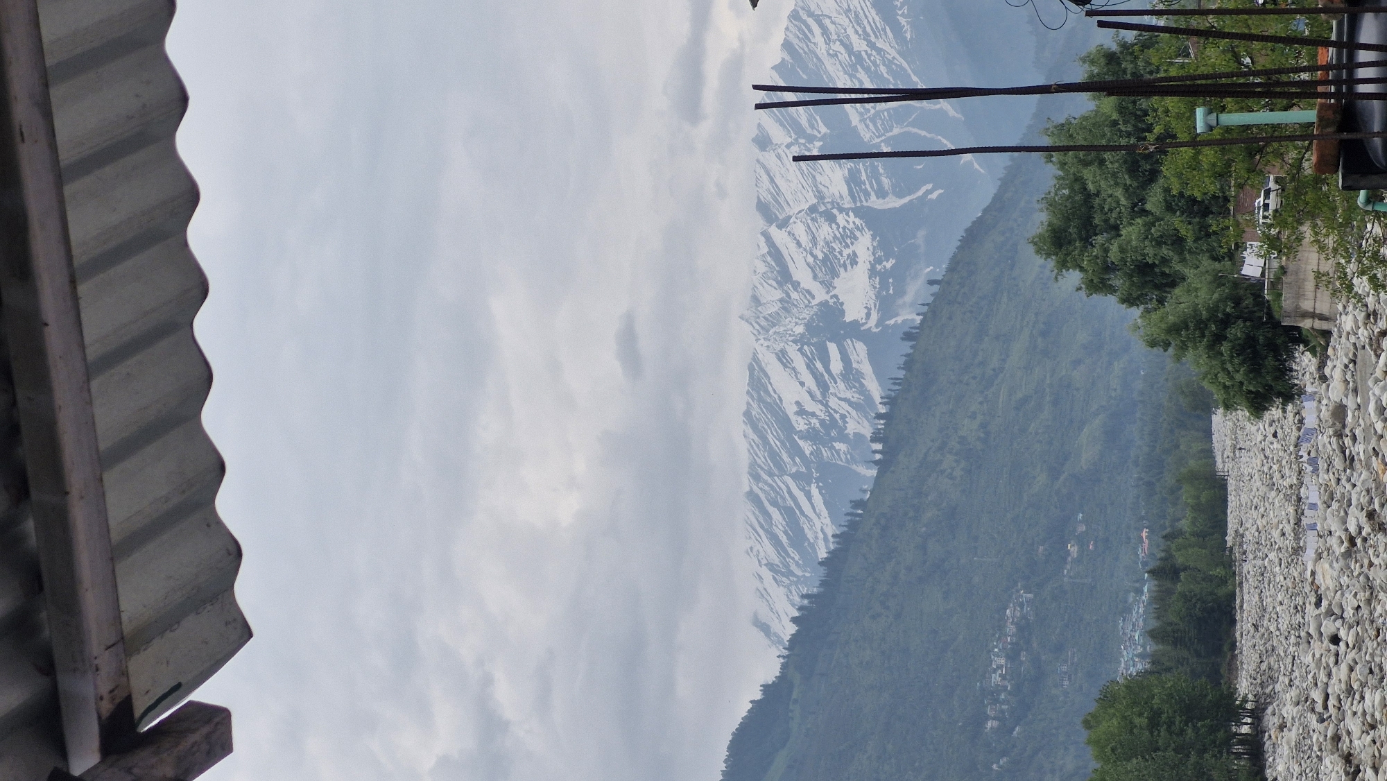 The image depicts a scenic view of a valley with snow-capped mountains in the background, likely in Vashisht, India. In the foreground, a dry riverbed filled with large, smooth stones stretches across the scene. A few people are visible scattered amongst the stones, and laundry is hung out to dry on lines. Lush green trees line the riverbed and the lower slopes of the mountains, which are dotted with small villages. The sky is overcast with gray clouds. A corrugated metal roof juts into the frame from the top, obscuring the very top of the sky. Some dark, vertical poles are present in the right side of the frame.