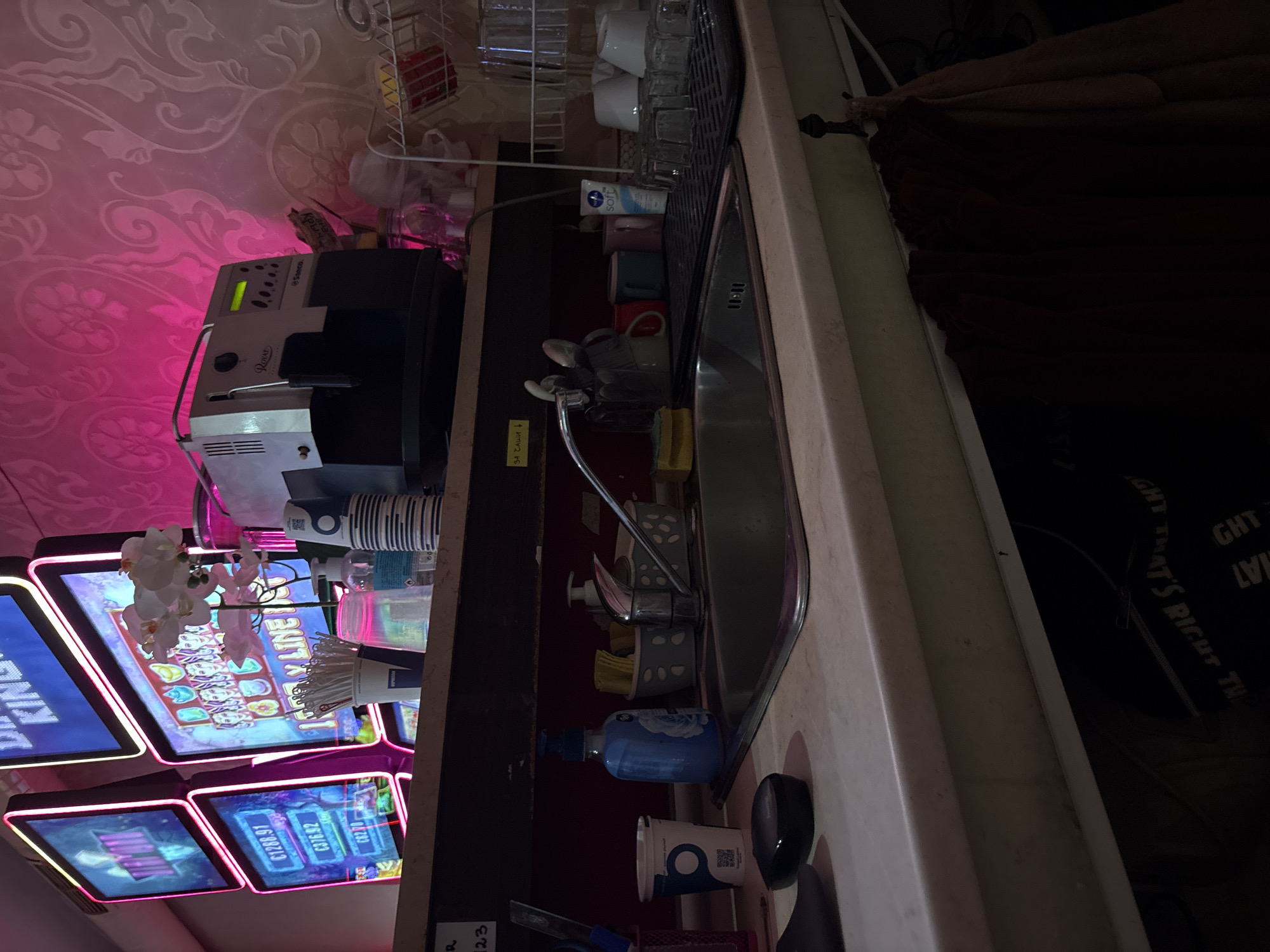 The image depicts a domestic scene, likely a kitchen or kitchenette, with elements suggesting a small business or personal living space. Prominently featured is a sink area with a faucet, soap dispenser, and cleaning supplies. To the left, several slot machine screens display colorful graphics and numbers, indicating a gambling establishment or a room with entertainment machines. A coffee machine is visible on a shelf above the counter. The overall impression is one of a functional, albeit slightly cluttered, space that combines elements of leisure and utility. The scene does not provide any specific visual cues to pinpoint its location in Karlovo, Bulgaria.