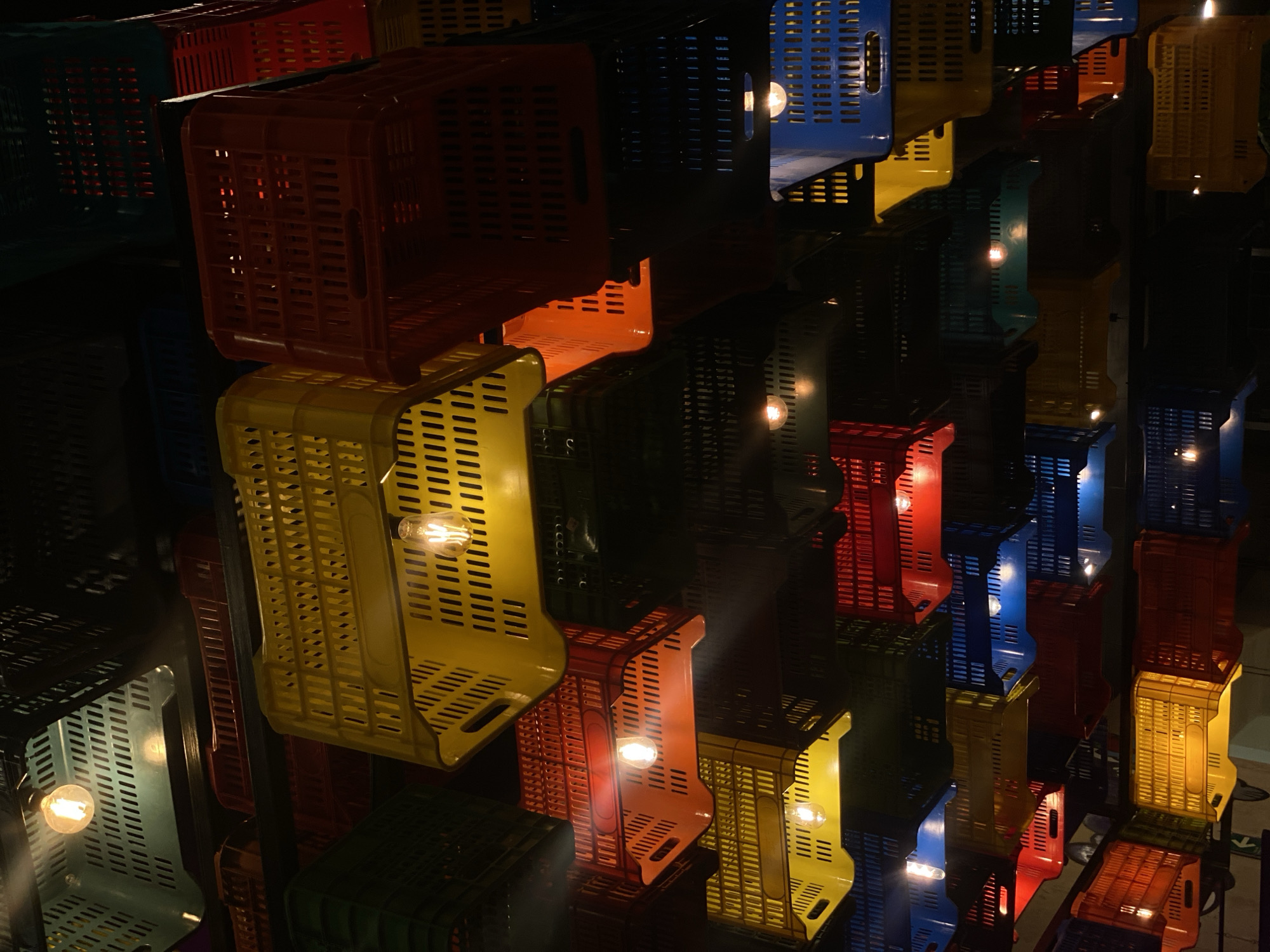 The image shows a creative lighting installation made of stacked, colorful plastic crates, each containing a light bulb. The crates vary in color, including blue, yellow, red, green, and orange. The lighting design seems to be suspended from the ceiling or mounted on a wall. The location appears to be in an urban setting.