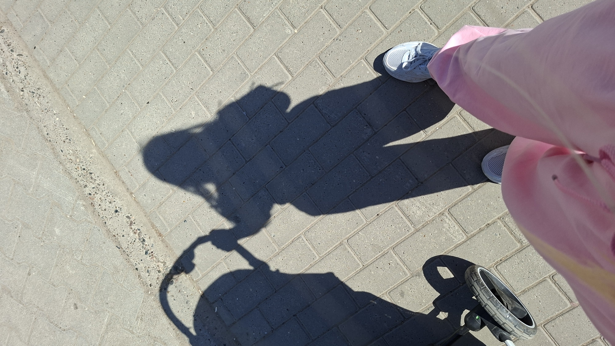 A person wearing pink pants and grey sneakers is standing on a grey brick sidewalk in Chaikovsky, Russia. The shadow of the person, a baby in a stroller, and the stroller itself are visible on the sidewalk.