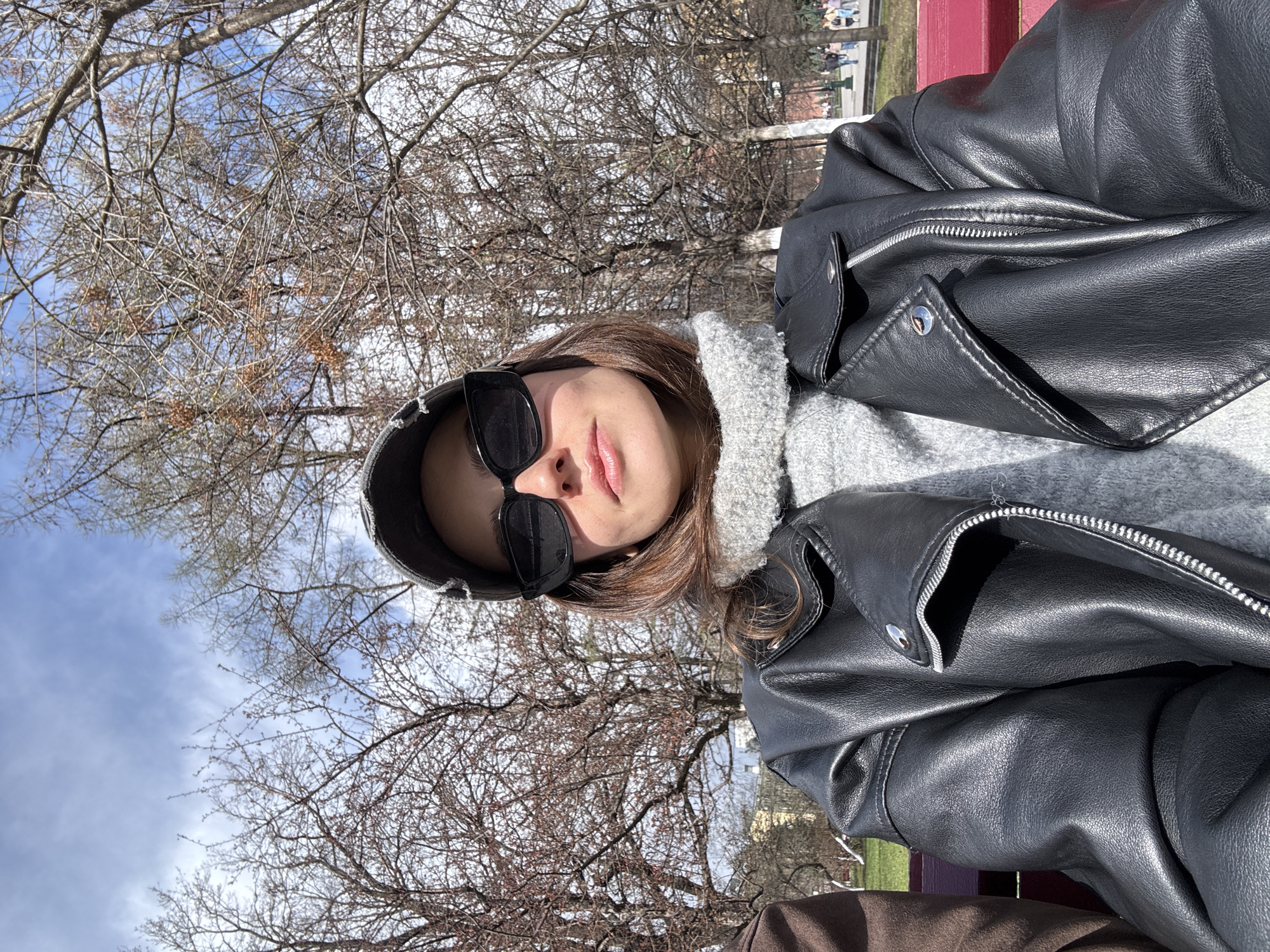 A young woman wearing a black leather jacket, gray sweater, and black sunglasses is taking a selfie outdoors. The background shows bare trees, a blue sky, and hints of buildings, suggesting an urban park setting. Based on the visual cues, the location can be pinpointed as Moscow, Russia.