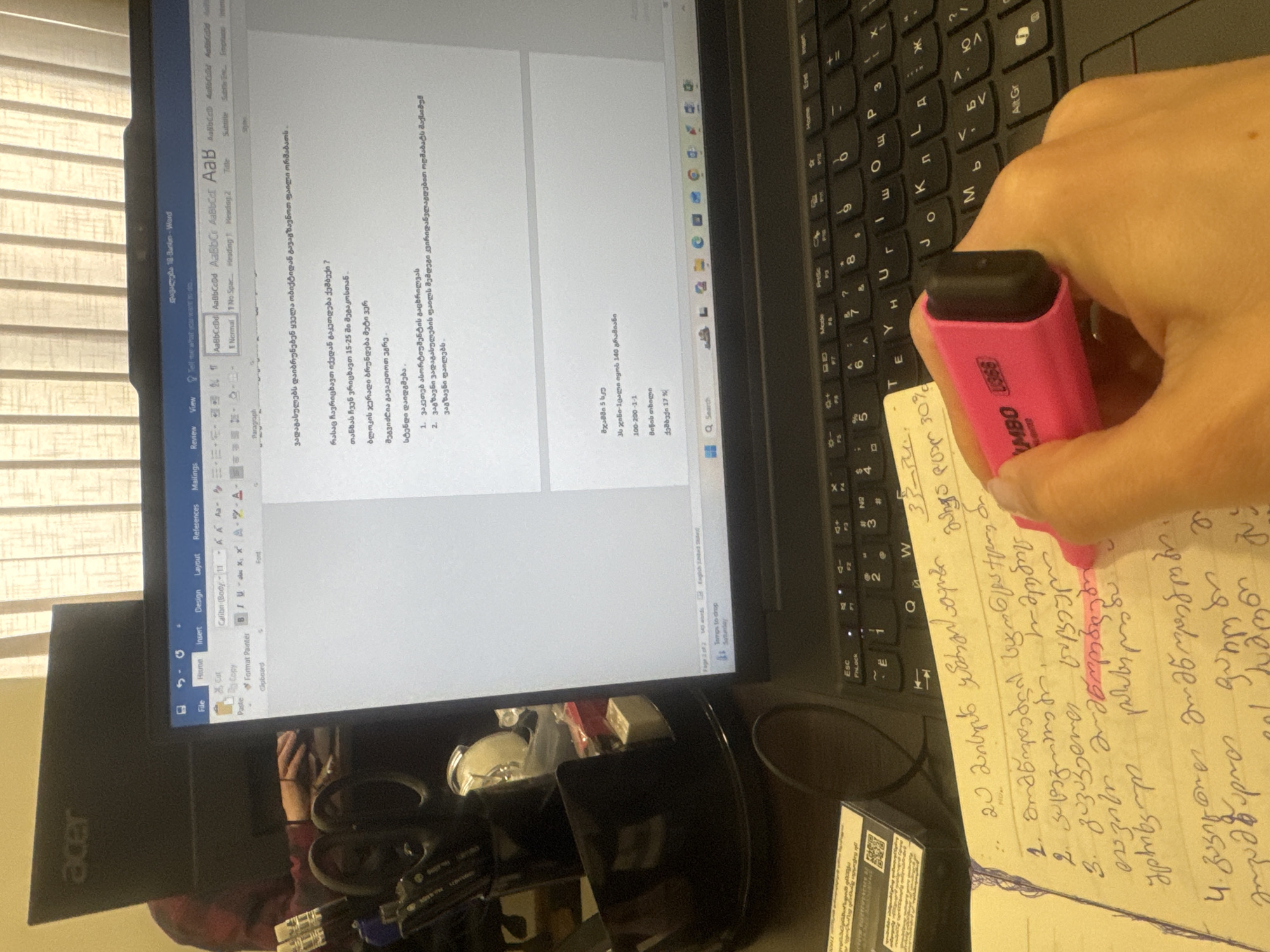 The image shows a person's hand holding a pink highlighter over a handwritten page filled with text in the Georgian language. The page appears to be notes taken in relation to a document open on a laptop screen nearby. The laptop screen displays a Microsoft Word document, also containing Georgian text. A keyboard is visible next to the notes and the laptop. The monitor of the computer is an Acer brand. The setting is a workspace, likely a home office, as indicated by the presence of office supplies. There are no clues in the photo that indicate the specific location in the city of Tbilisi, Georgia.