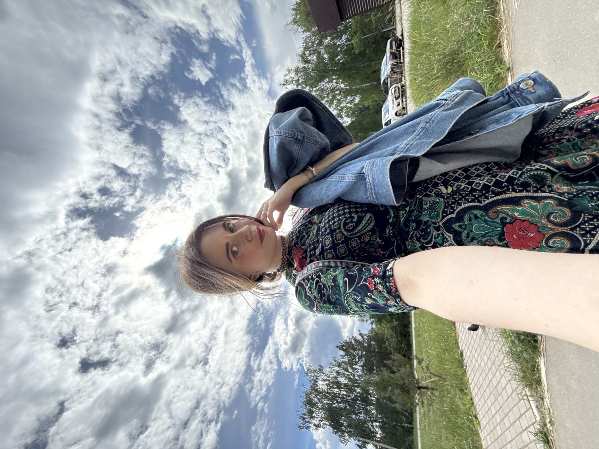 A portrait-oriented, low-angle shot features a woman outdoors against a backdrop of a dramatic sky with white clouds and blue patches. The woman, positioned in the left half of the frame, is wearing a dark, patterned dress with red and green floral motifs and a black choker. A light blue denim jacket is draped over her left shoulder and arm. Her right arm is extended towards the camera. She has light brown hair, fair skin, and is wearing red lipstick. Her expression is neutral, with her gaze directed towards the camera.
To the right of the woman, a paved pathway runs parallel to a grassy area. Beyond the grass, several vehicles are parked. In the upper right corner, the edge of a building is visible. The lighting suggests it is daytime, likely mid-morning or afternoon, with the sun casting shadows and illuminating the clouds. The overall atmosphere is casual and relaxed. There is no visible text in the image.