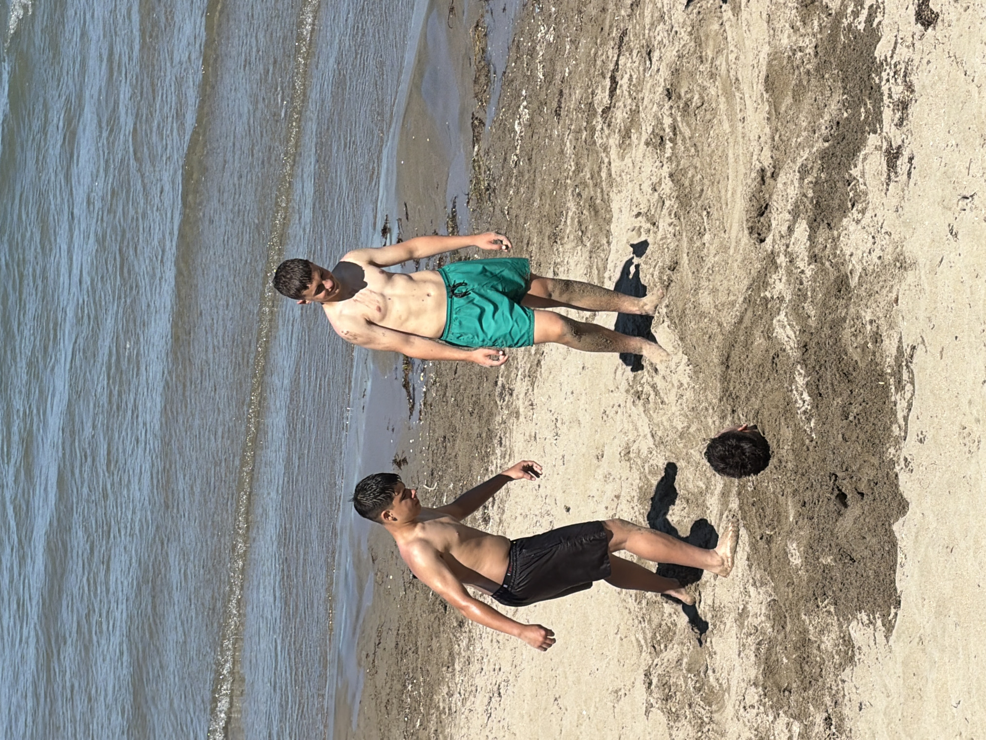 Two young males are on a sandy beach near the sea. One male, wearing black shorts, is on the left, facing a mound of sand. The other male, wearing green shorts, is on the right, looking down. A person's head is visible emerging from the mound of sand. The environment is a beach with calm water in the background. The scene appears to be located in Tsarevo, Bulgaria.