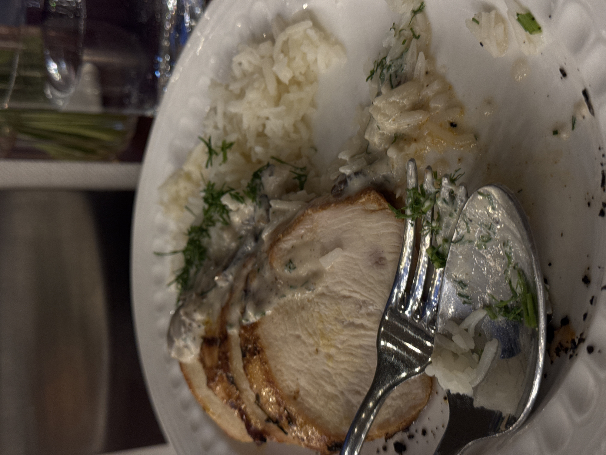 The image shows a partially eaten meal on a white plate. The main component appears to be sliced meat, likely turkey or chicken, covered in a cream sauce with mushrooms and dill. Next to the meat is a portion of white rice. A silver fork and spoon rest on the plate, partially covered in sauce and rice. The overall setting seems to be a domestic environment, but there's nothing to specifically indicate that the photograph was taken in Yerevan, Armenia.
