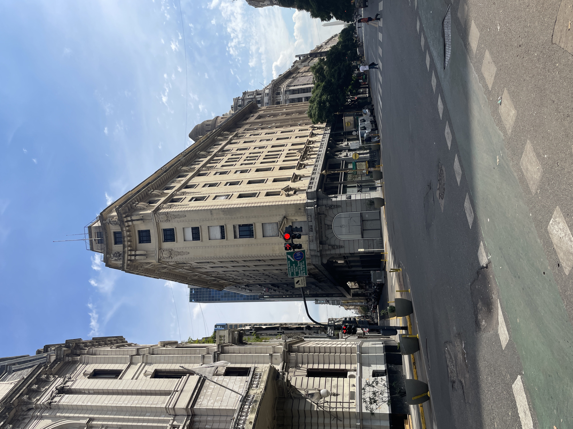 The photo shows a street view in Buenos Aires, Argentina. The most prominent structure is the Edificio Sud América, a wedge-shaped building with a beige facade and repetitive window patterns. A traffic light on the building displays a red signal. The building stands at the intersection of Diagonal Norte and Calle Florida. The street is paved with asphalt and marked with a green bike lane. Concrete bollards line the edge of the road. Pedestrians and vehicles are visible in the distance. The sky is partly cloudy. The architectural style of the surrounding buildings is consistent with the historic center of Buenos Aires.