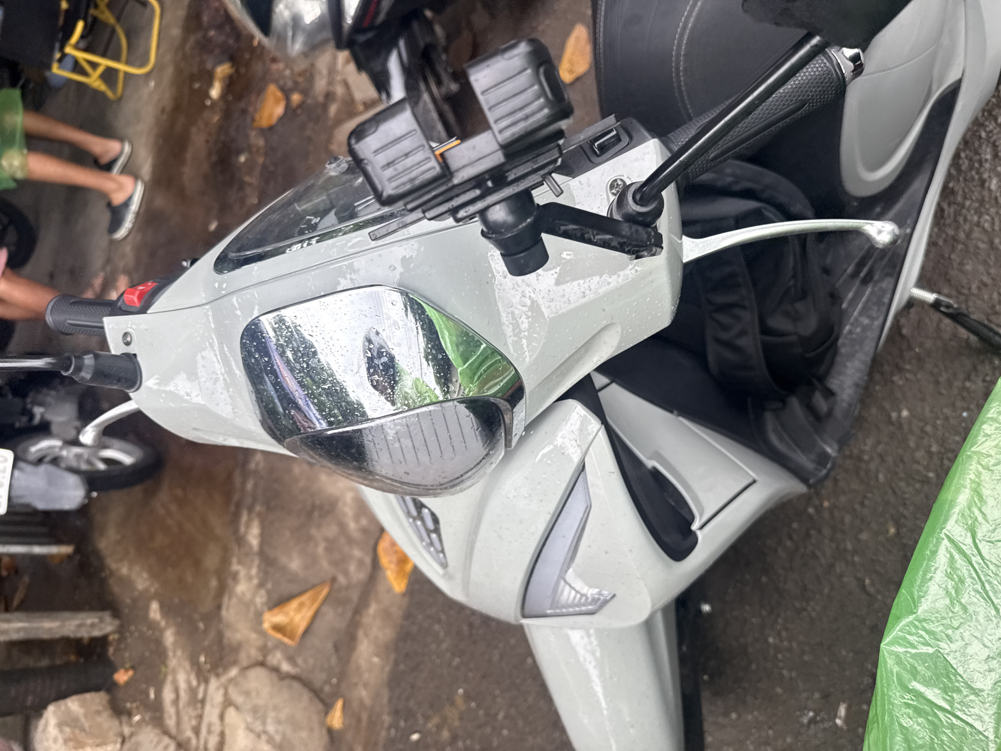 The image captures a close-up, top-down view of a light gray scooter parked outdoors, likely in Nha Trang, Vietnam, suggested by the presence of several conical brown leaves scattered on the wet, paved ground. The scooter's surface is speckled with water droplets, indicating recent rain. A black phone mount is attached to the handlebars. In the background, blurred figures of people are visible, one wearing dark shoes and the other standing barefoot, with another person wearing a green poncho. A yellow metal chair is also present. The overall environment appears to be a street or walkway, with cracked concrete and damp, muddy patches.