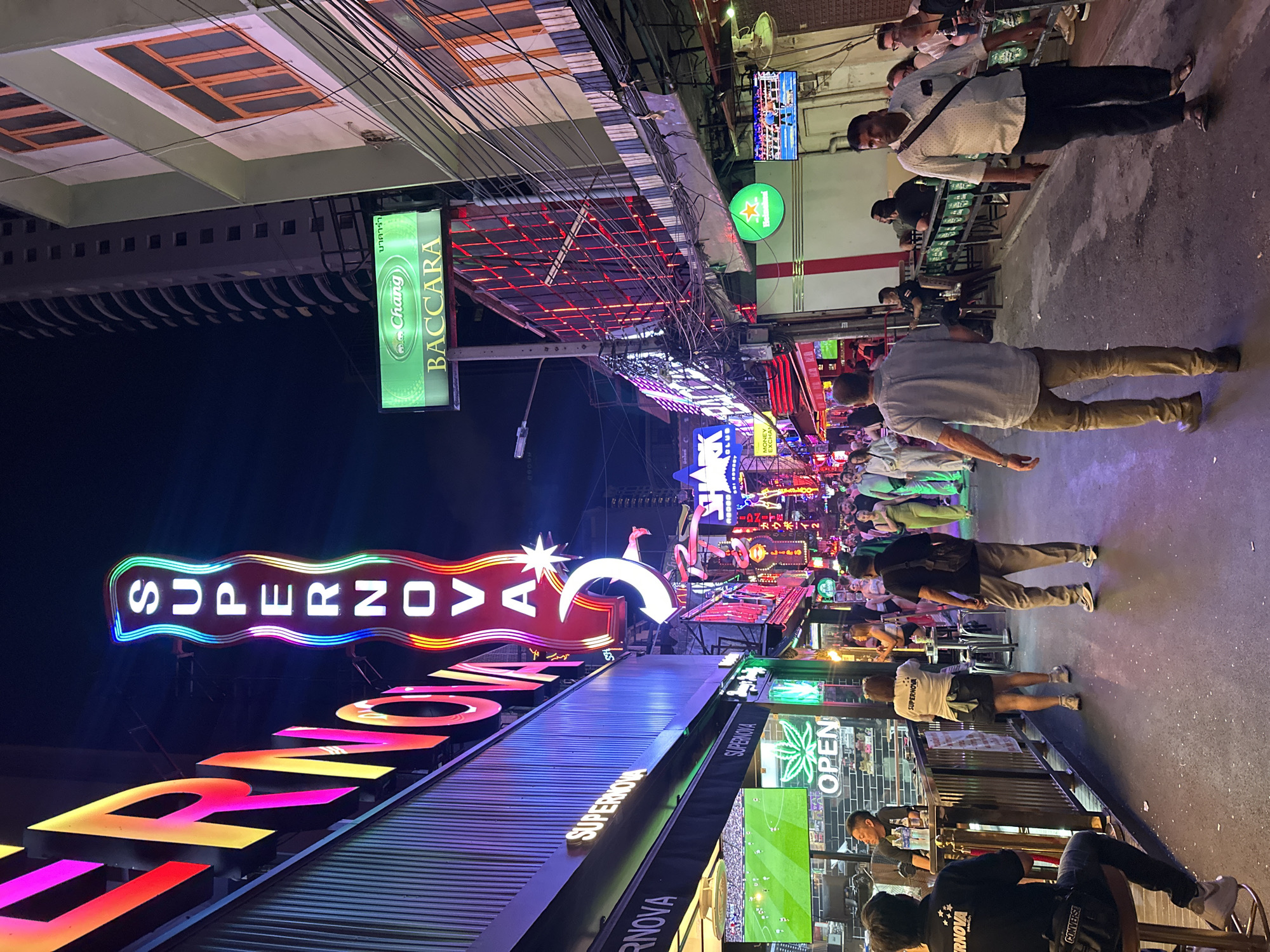 The image depicts a street scene at night in Bangkok, Thailand. The street is filled with pedestrians and is lined with businesses adorned with neon signs. Prominently displayed is a large, colorful neon sign that reads "SUPERNOVA." A sign above it reads "RNOVA".
There are people walking along the street and seated at tables outside the businesses. The lighting is bright and vibrant due to the neon signs and streetlights. Some of the people have fair skin. There is also a large green sign reading "Chang BACCARA". The photo is taken in a slightly skewed perspective. Over all of the shops and signage there are large webs of cables.