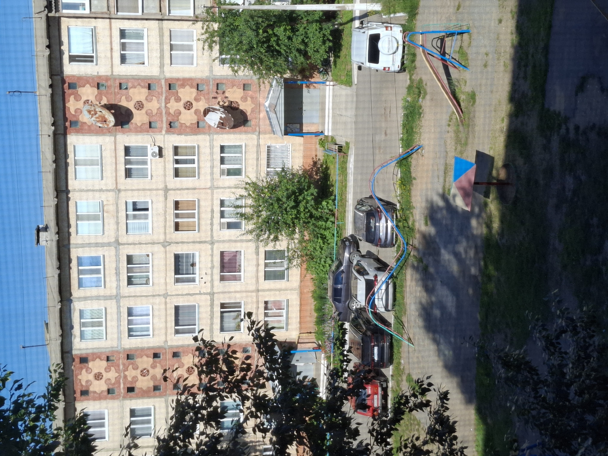 This image depicts an outdoor scene in the city of Jitiqara, Kazakhstan. In the foreground, there is a playground area with a colorful umbrella-like shade structure and a slide with a metal frame. Grass and patches of dirt surround the playground equipment.
Dominating the middle ground is a parking lot with several cars parked. Visible vehicles include a red sedan, a black SUV, a white SUV with the word "PATRIOT" on the back, and a dark-colored sedan. Some of these cars are partially obscured by foliage. Colorful playground safety barriers, consisting of blue, green, and red poles with wires, are present between the parking area and the playground.
The background features a multi-story apartment building. The building has a light-colored facade with decorative elements, including geometric patterns and circular motifs in shades of pink and beige. Several windows with white frames are visible on the building's exterior. Two satellite dishes are attached to the building's facade, positioned vertically. An awning with blue supports covers the entrance to the building. Trees with green foliage are situated to the left and right of the building entrance, and also along the building's facade. The sky above is clear and blue.