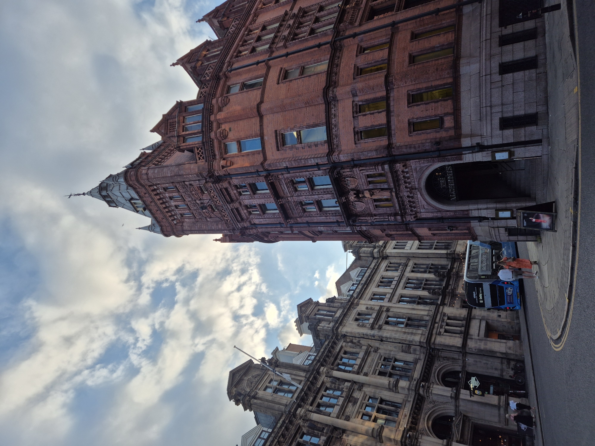 This is a low-angle, eye-level, wide shot of a street in Nottingham, United Kingdom, featuring two distinct architectural styles of buildings. The building on the right is a grand, reddish-brown brick structure with ornate detailing and a prominent steeple, appearing to be a historical building. An arched entrance reads "THE ALCHEMIST". The building on the left is a lighter-colored stone structure, also appearing historical, with multiple windows and decorative elements. A blue double-decker bus is visible on the street in front of the stone building. Two individuals, a man and a woman, are walking away from the camera on the sidewalk between the bus and the brick building. The sky above is partly cloudy with patches of blue. The overall scene suggests a bustling urban environment with a blend of historic architecture.