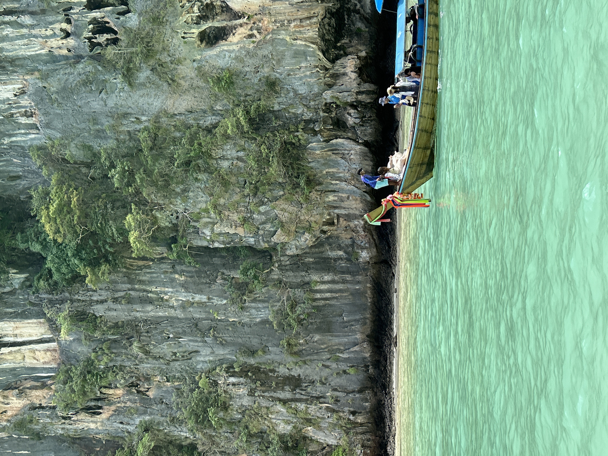 In Ao Nang, Thailand, a longtail boat with several people on board navigates through the turquoise waters of the Andaman Sea, approaching a towering limestone cliff face covered in dense vegetation. The cliff dominates the background, its rugged texture and dark crevices suggesting a natural and ancient landscape. The water is clear and calm, with subtle ripples disturbing its surface. On the boat, a man in a blue shirt stands near the bow, while other passengers, including women in light-colored clothing, are seated further back. The boat itself is classic longtail design, with a distinctive colorful streamer attached to its prow. The scene evokes a sense of tropical adventure and exploration.