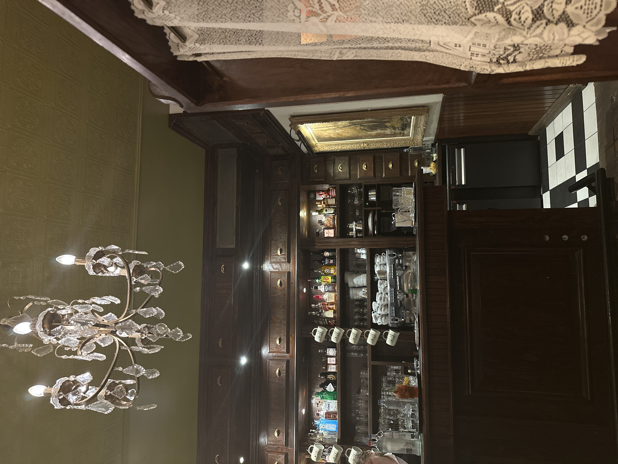 The image depicts the interior of a dimly lit, traditional bar in Bruges, Belgium. The scene is dominated by dark, polished wood paneling and cabinetry, with a prominent bar counter in the foreground. Above the bar, shelves are stocked with various bottles of alcohol, glassware, and ceramic mugs hanging from hooks. An espresso machine is visible on the counter. A crystal chandelier hangs from the textured, olive-green ceiling, casting a warm glow. To the right, a framed painting hangs on the wall, partially obscured by delicate white lace curtains. The floor features a black and white tiled pattern. A person, partially visible at the far left, appears to be a bartender or server. The overall ambiance suggests an old-world European establishment.