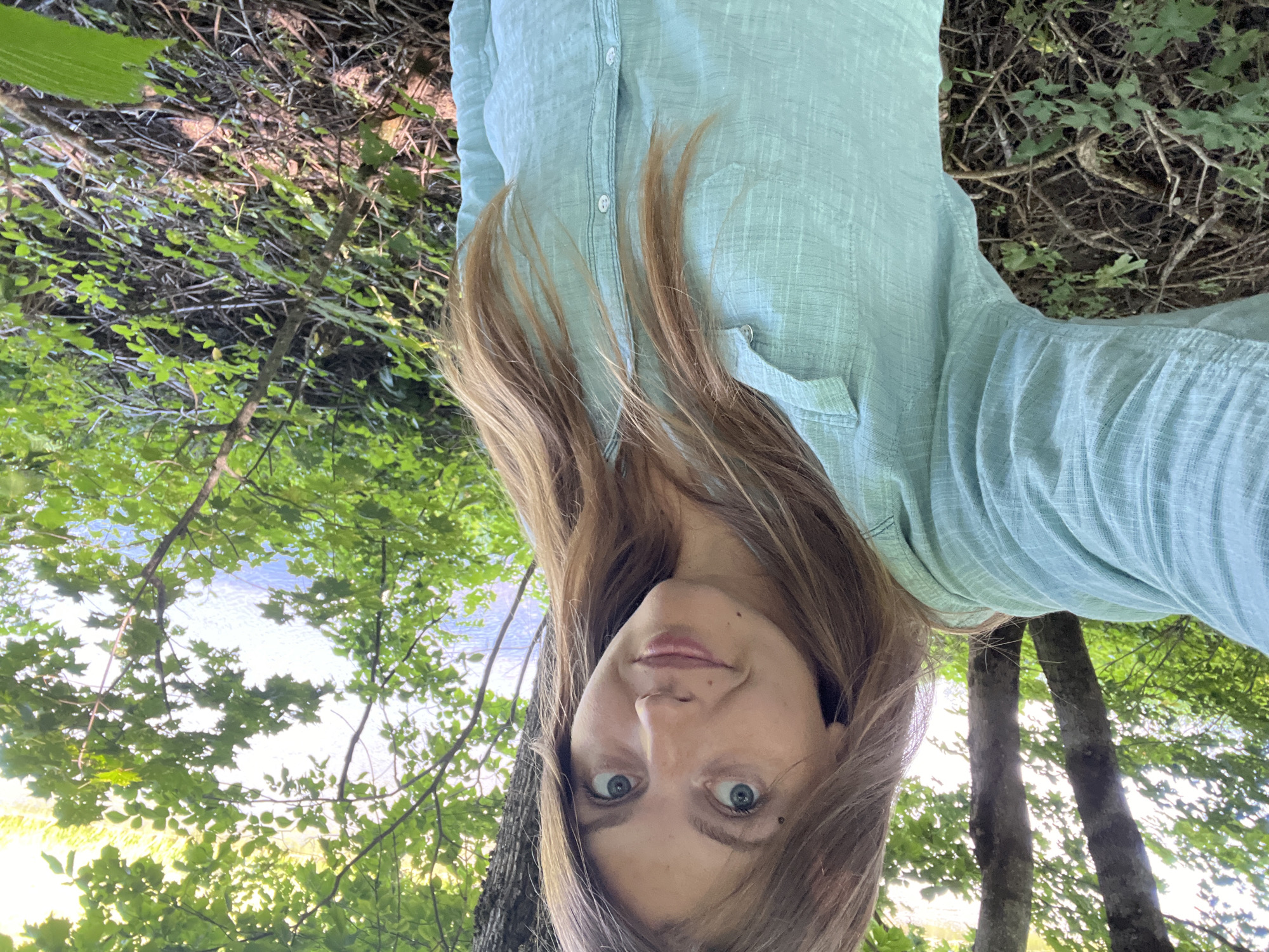 A woman with long, light brown hair takes a selfie in a wooded area near a body of water. She is wearing a light blue, long-sleeved button-down shirt. The background consists of trees, green foliage, and the glinting surface of water, suggesting a natural, outdoor setting. The woman's expression is casual and direct towards the camera. The scene appears to be in a park or a natural reserve, potentially within the vicinity of Германавіцкі сельскі Савет, Belarus, given the vegetation and water.