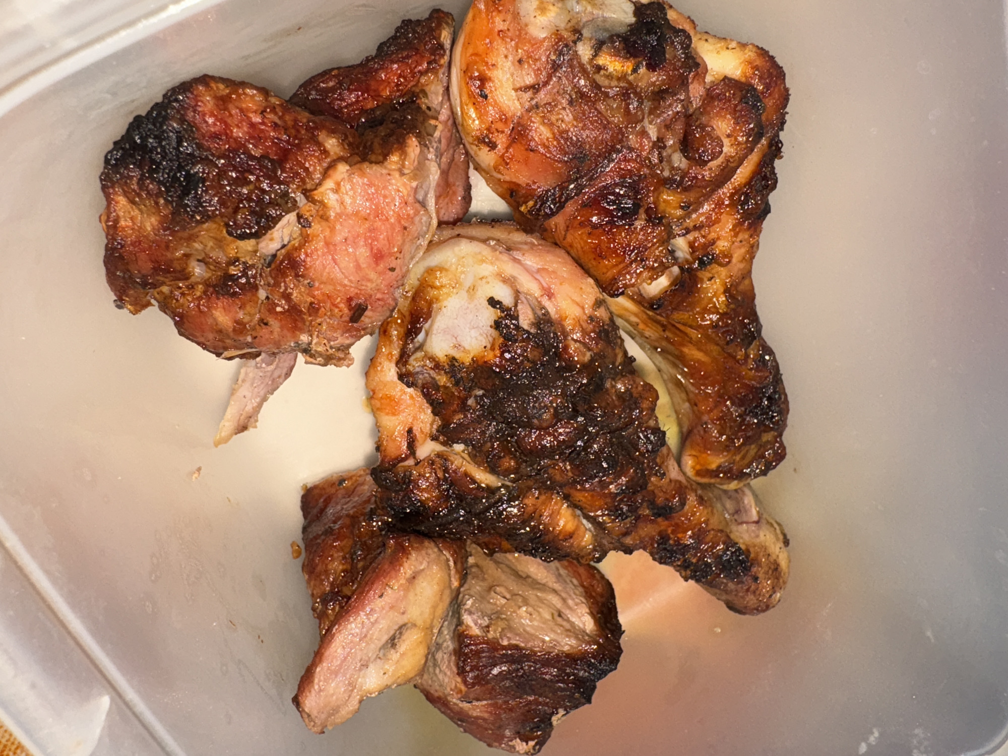 The image shows four pieces of cooked chicken in a plastic container. The chicken appears to be grilled or roasted, with some charring. The container is translucent and shows the chicken in close-up detail. The location in the city of Rusalevka, Russia is not determinable from the image. No people, actions, or interactions are visible. The environment is limited to the inside of the container.