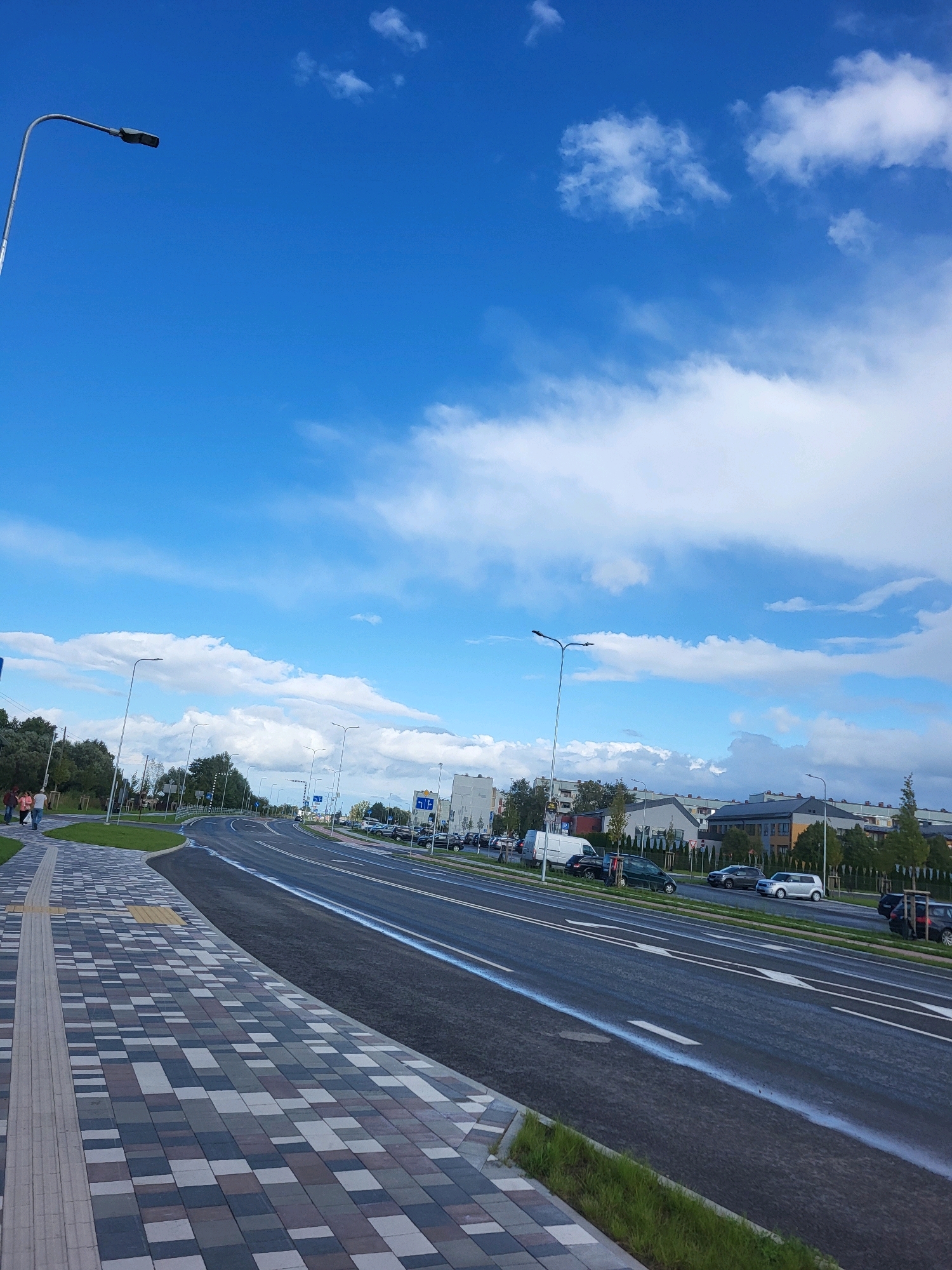 The image captures a street scene in Jelgava, Latvia, under a bright blue sky with scattered white clouds. A wide, paved road curves to the right, with multiple lanes marked by white arrows. On the left, a patterned sidewalk with tactile paving runs alongside the road. Several streetlights are visible along the road. In the background, there are trees and residential buildings. A few cars are parked or driving on the road. Two individuals are walking on the sidewalk. The scene appears to be a contemporary urban environment, possibly a newer development or a recently renovated street.