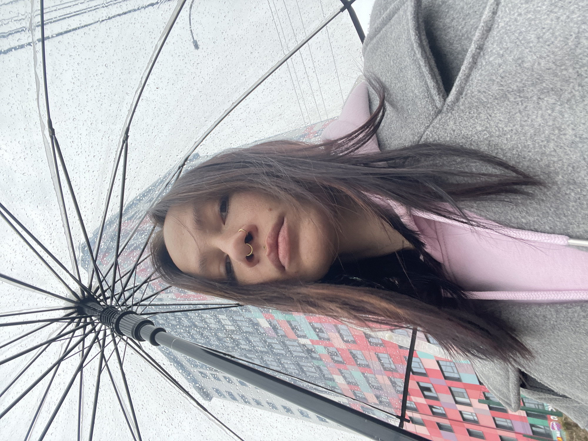 A person with dark hair and a nose ring is seen from a low angle, holding a clear umbrella covered in raindrops. The person is wearing a pink hoodie under a grey coat. The background shows a multi-colored building with a pattern of windows and red and purple shapes; this building is located in Izhevsk, Russia.