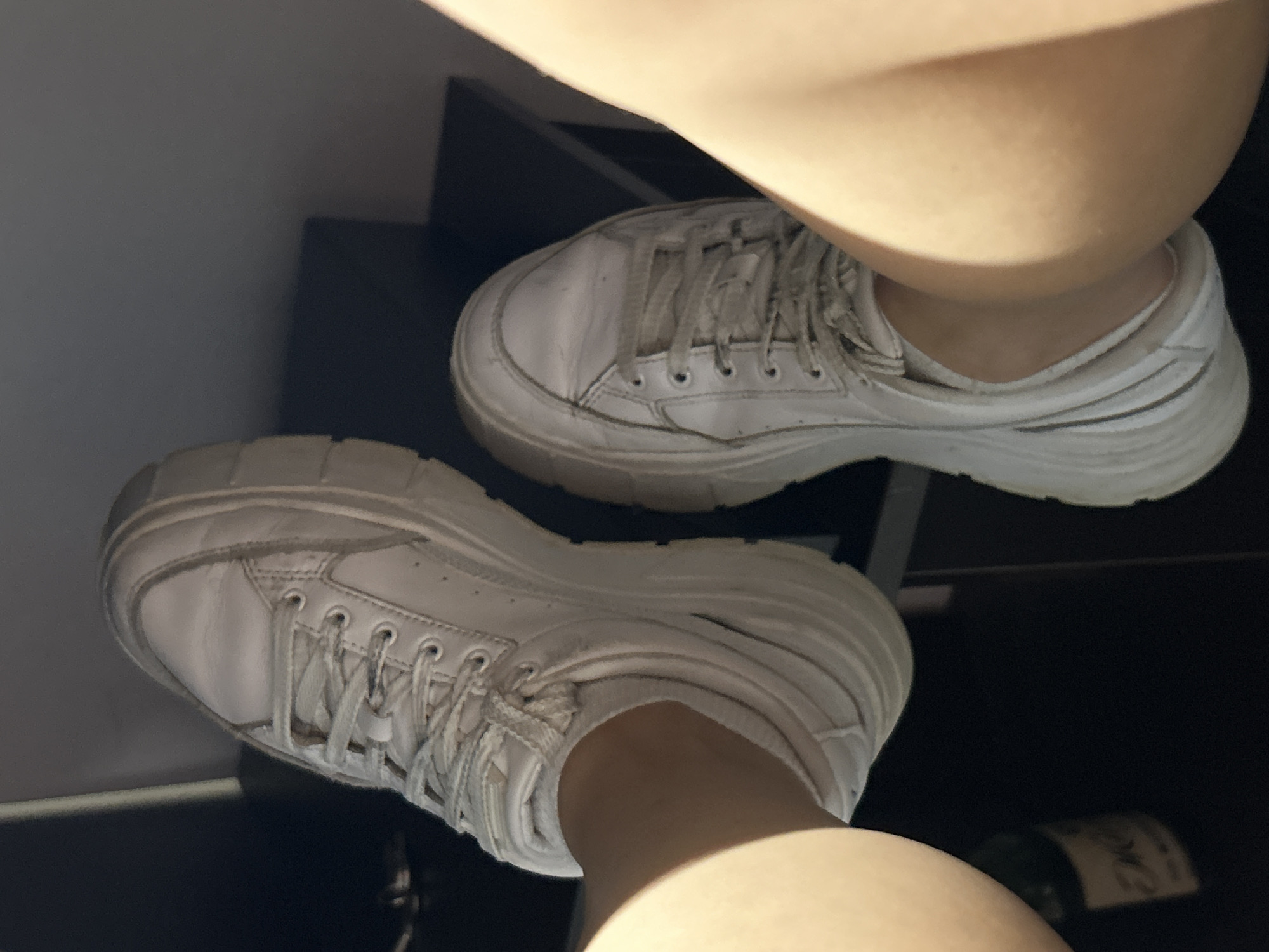 Two white platform sneakers are visible in the image. The left sneaker is closer to the viewer and appears to be made of leather. It has white laces and a thick, white sole. The right sneaker is partially visible behind the left one and appears to be identical in style. Both shoes show signs of wear, with some creasing and scuff marks. A person's legs and feet are wearing the shoes. The background is dark and out of focus, but appears to be an indoor setting with some dark furniture. A small portion of a green bottle with a label is visible in the bottom left corner. The lighting suggests it may be daytime with sunlight casting shadows. The location is identified as Tbilisi, Georgia.