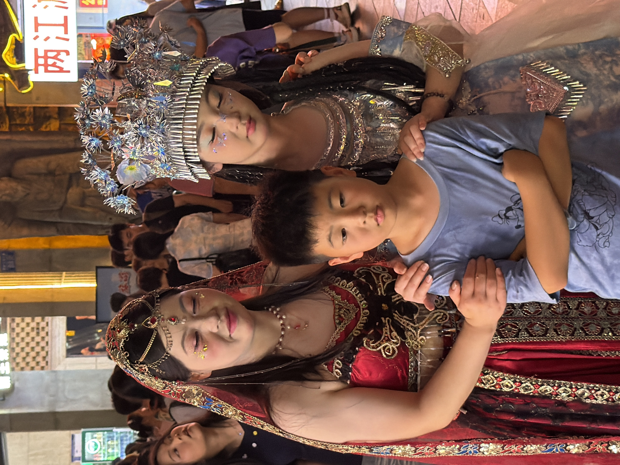 The image shows a group of people in what appears to be an outdoor setting at night, possibly on a street in Yuzhong District, China. The main subjects are two young girls and a young boy, all dressed in elaborate costumes.
The girl in the foreground is wearing a traditional Indian-inspired outfit in red and gold, adorned with intricate embroidery, beads, and a bejeweled headpiece. She has glitter on her face and her eyes are closed, suggesting a posed or performance moment. A pearl necklace is visible around her neck.
Behind her, a younger girl is wearing a silver, star-themed headdress and a shimmering, sequined costume. She also has glitter on her face and appears to be looking down.
A young boy, wearing a light blue t-shirt with a cartoon character on it, is positioned between the two girls, his arm around the girl in the red outfit. He is looking towards the camera.
The background is blurred, but appears to be a street scene with some lights and structures. There is a vertical sign with Chinese characters on it on the left side of the image. The lighting suggests it is nighttime. The overall scene seems to be a celebration or a cultural event, possibly a performance or a parade. The expressions on the faces of the children are calm, and the overall mood is festive.