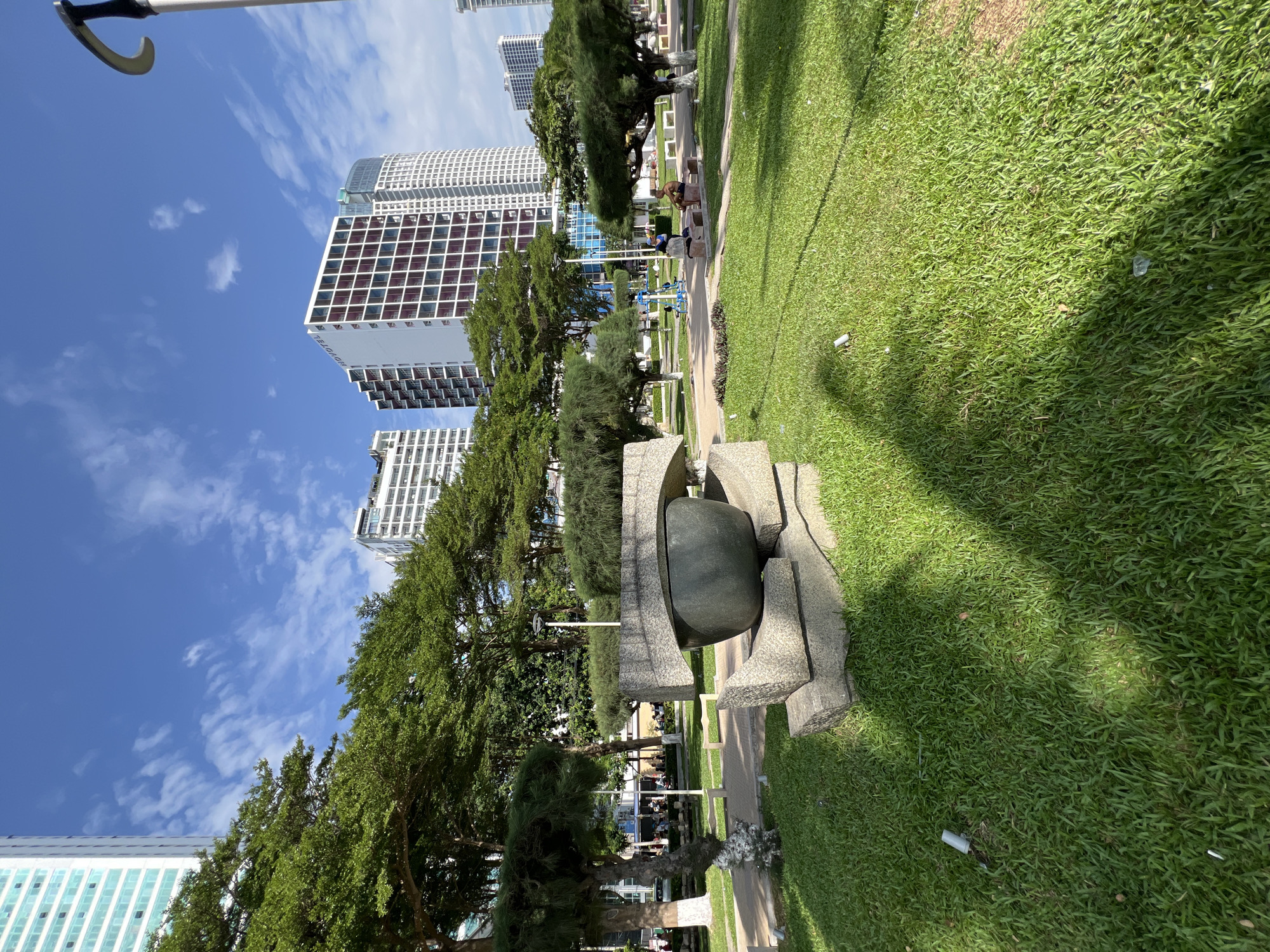 This image captures a scene in a public park located in the city of Nha Trang, Vietnam, likely near the beachfront. The foreground is dominated by a well-maintained green lawn with a shadow cast from a sculpted object. This sculpture appears to be made of rough-hewn stone blocks, with a large, dark, bowl-shaped element resting on top. Behind the sculpture, a paved walkway curves through the park, lined with manicured trees that have a distinct, umbrella-like canopy. In the mid-ground, several people are visible. Two individuals are seated on a park bench, and further back, more people can be seen gathered near what looks like exercise equipment. The background of the image is characterized by tall, modern buildings, indicative of urban development along the coast. The sky is a vibrant blue with scattered white clouds, suggesting fair weather. The overall environment is clean and orderly, with a blend of natural landscaping and urban architecture.