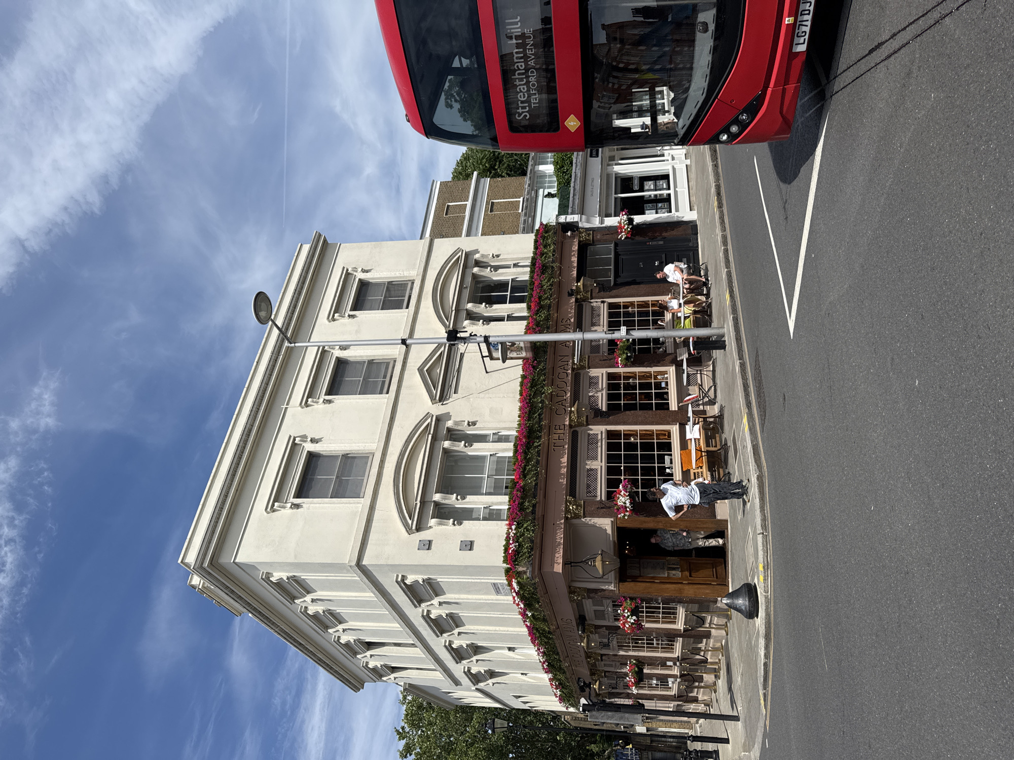 A sunny, daytime, eye-level, exterior shot captures the corner of "The Cadogan Arms" pub and a red double-decker bus at an intersection in Chelsea, London, United Kingdom. The pub, a cream-colored building with ornate detailing and a prominent flower box along its upper facade, features large windows and an open doorway with two men visible inside. Outside the pub, tables and chairs are set up on the pavement, with two individuals seated at one table. The bus, a modern double-decker, is partially visible on the right side of the frame, with its destination displayed as "Streatham Hill Telford Avenue". The street is paved, and a streetlamp stands near the pub. The sky is blue with scattered white clouds.