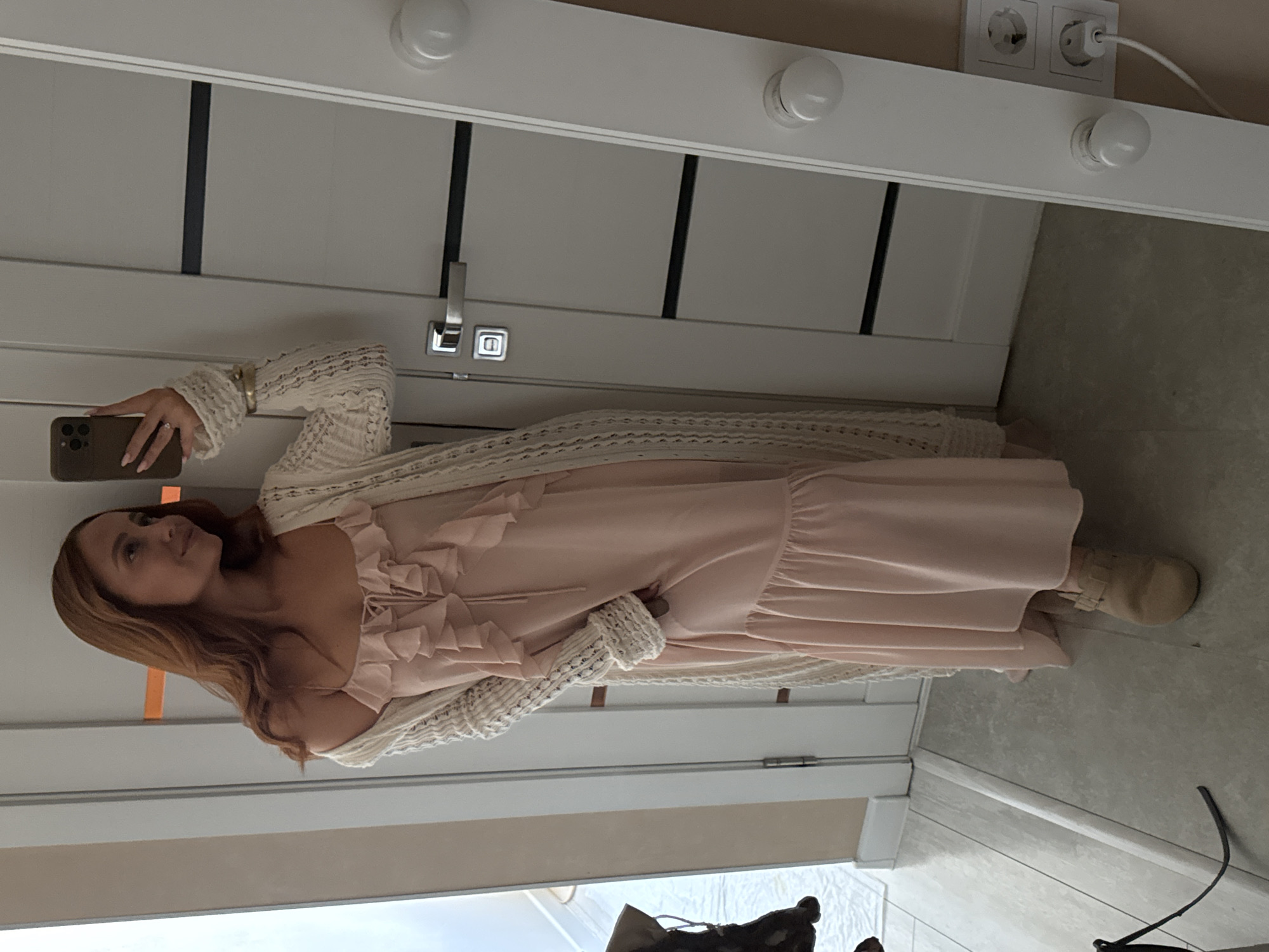 A woman with long, wavy, reddish-brown hair stands in a room, taking a mirror selfie with her phone. She is wearing a pale pink, ruffled maxi dress and a cream-colored, open-knit cardigan. Her right hand is raised, holding the phone, and her left hand is tucked into the side of her dress. She is wearing tan clogs. The background shows a white door with black horizontal accents, a silver door handle, and a white vanity mirror with light bulbs along the side. Electrical outlets are visible on the wall to the right of the mirror. The floor is tiled with light-colored wood-grain planks. The lighting is soft and natural. The scene appears to be in a residential interior, likely a bedroom or dressing area. There is no identifiable information suggesting the location is in Chaikovsky, Russia.
