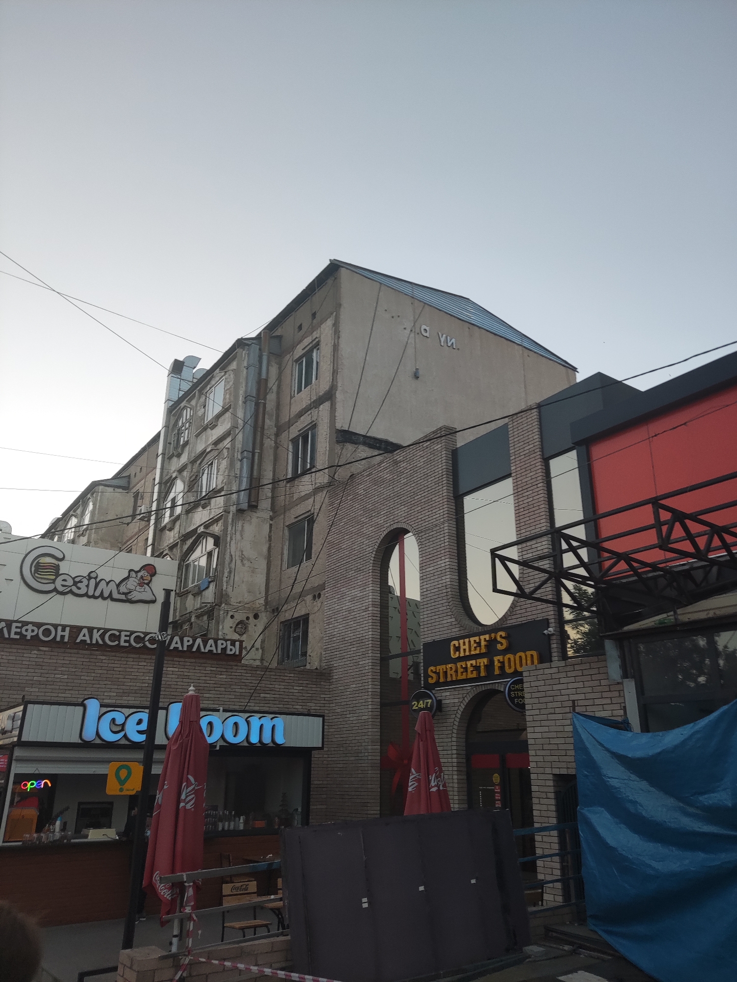 The image shows a street scene in Shymkent, Kazakhstan, with a focus on storefronts. The time of day appears to be late afternoon or early evening, given the soft light. An older, weathered apartment building with multiple windows occupies the left side of the frame, contrasting with a more modern, brick-faced building to its right.
Several businesses are visible. On the left, a sign with "Сезім" (Sezim) and a cartoon of a hamburger and a chicken is illuminated. Below it, text in Cyrillic reads "ЛЕФОН АКСЕССУАРЛАРЫ" (Telephone Accessories). Adjacent to this is a brightly lit sign for "Ice Dom." A red umbrella with branding is partially visible in front of the Ice Dom establishment.
To the right, the brick-faced building features signage for "CHEF'S STREET FOOD" with a "24/7" symbol. The building has large windows and a red accent section. A blue tarp is draped over some construction or debris in the foreground on the right. The sky above is clear and pale. The overall impression is a bustling urban environment with a mix of old and new architecture and commercial activity.