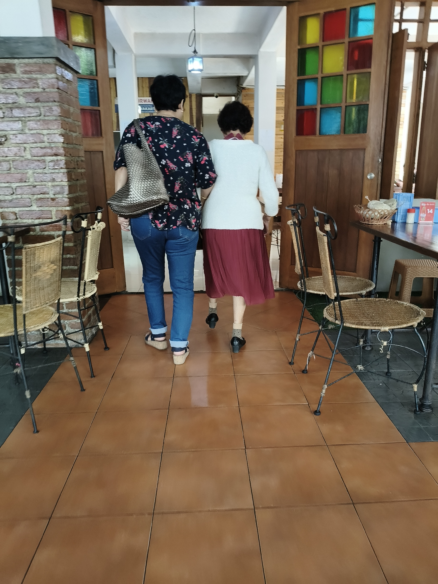 Two women are walking away from the camera through an indoor walkway with tiled flooring. The woman on the left has short black hair and wears a dark floral patterned shirt, blue jeans, and sandals. She carries a woven tote bag over her left shoulder. The woman on the right has short black hair and wears a white sweater, a red pleated skirt, and black heels. They are walking towards an open doorway with a wooden frame and stained glass panels in various colors. To the left of the doorway is a brick wall. In the foreground and to the left are two wicker chairs with black metal frames. To the right of the doorway, a wooden table is visible with chairs and a basket. The scene appears to be in a cafe or restaurant in Bandung City, Indonesia, suggested by the architectural style and cultural context.