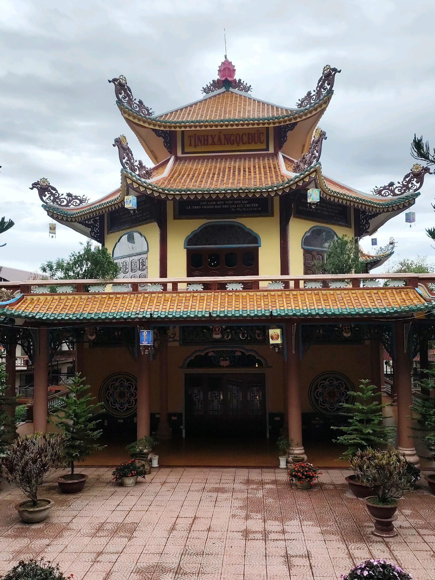 The image shows the entrance of a Buddhist temple in Da Lat, Vietnam, likely Truc Lam Zen Monastery or a similar structure given the architectural style. The temple features a multi-tiered, curved roof with ornate dragon-like carvings at the eaves. The building is painted in yellow and orange, with red accents on the roof tiles and decorative elements. A sign in Vietnamese, reading "TINH XANG NGOC DUC," is visible above the main entrance. The courtyard in front of the temple is paved with patterned tiles. Several potted plants and trees are arranged around the courtyard, including small evergreen trees and flowering bushes. The sky is overcast. There are no people visible in the image.