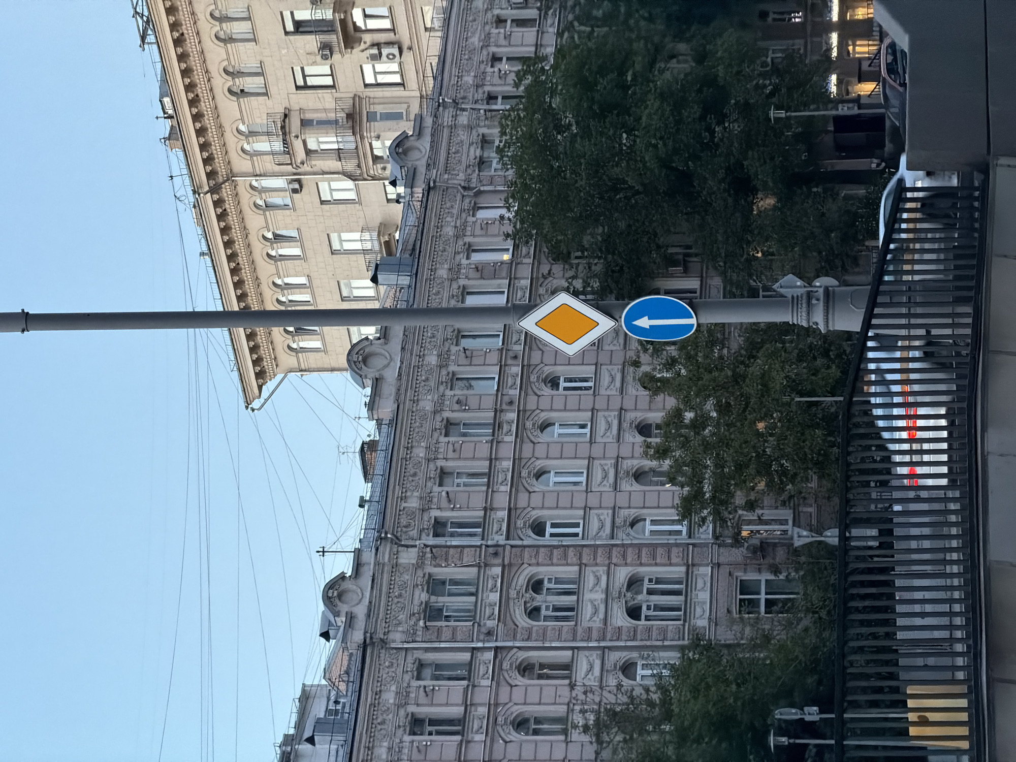 The image presents an urban street scene in Moscow, Russia, characterized by classical architecture and clear skies. In the foreground, a sturdy gray lamppost stands, holding two prominent traffic signs. The upper sign is a yellow diamond with a white border, signifying a priority road. Directly beneath it, a blue circular sign features a bold white arrow pointing to the left, indicating a mandatory turn or direction.
Behind the lamppost, two grand, multi-story buildings dominate the midground. The closest building exhibits elaborate architectural detailing, including ornate window frames, decorative pilasters, and cornices on its pale, possibly beige or light gray, facade. Its numerous windows vary in shape, with some being rectangular and others featuring elegant arched tops. Behind and slightly to the left, another structure of similar architectural style can be seen. Several lush green trees are scattered across the scene, partially obscuring the buildings.
The sky above is a clear, bright blue, with thin utility wires stretching across. To the far right, a dark metal fence or railing borders the scene, and beyond it, glimpses of artificial lighting suggest the approaching evening, possibly from streetlights or interior lights of buildings. No people or specific activities are visible, lending a quiet, contemplative atmosphere to the bustling urban environment. The image captures a blend of historical grandeur and everyday city elements under a calm sky.