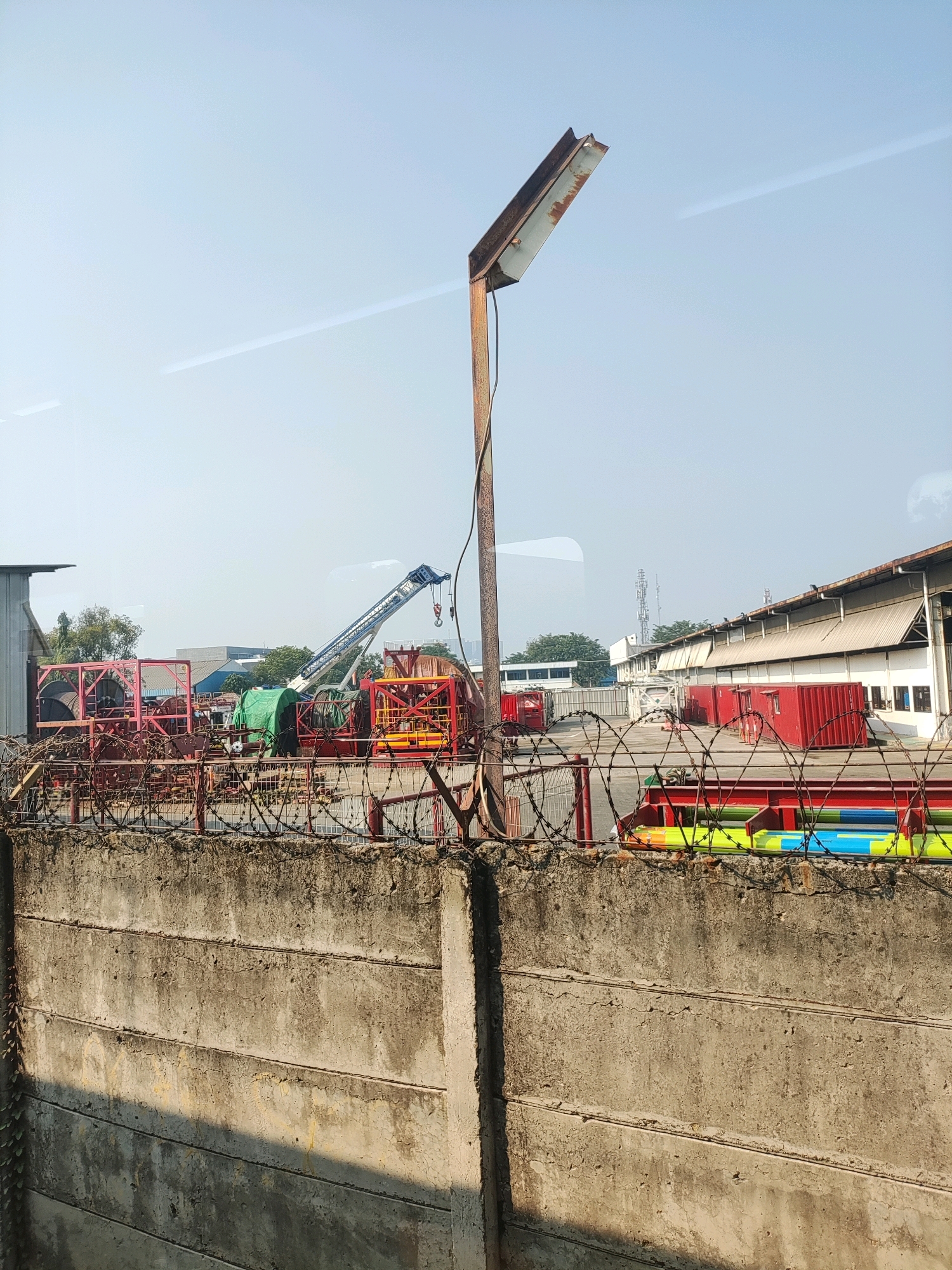 The image depicts an outdoor industrial area, likely a port or storage facility, in Bekasi, Indonesia. A concrete wall topped with barbed wire fence runs across the foreground. Behind the fence, several large industrial items are visible, including red metal cages containing spools of cable or wire, a green tarp covering some equipment, and a blue crane with its arm extended. In the background, there are industrial buildings, shipping containers, and communication towers under a hazy sky. A rusty metal lamppost stands prominently to the right of center. The scene appears to be an active industrial zone.